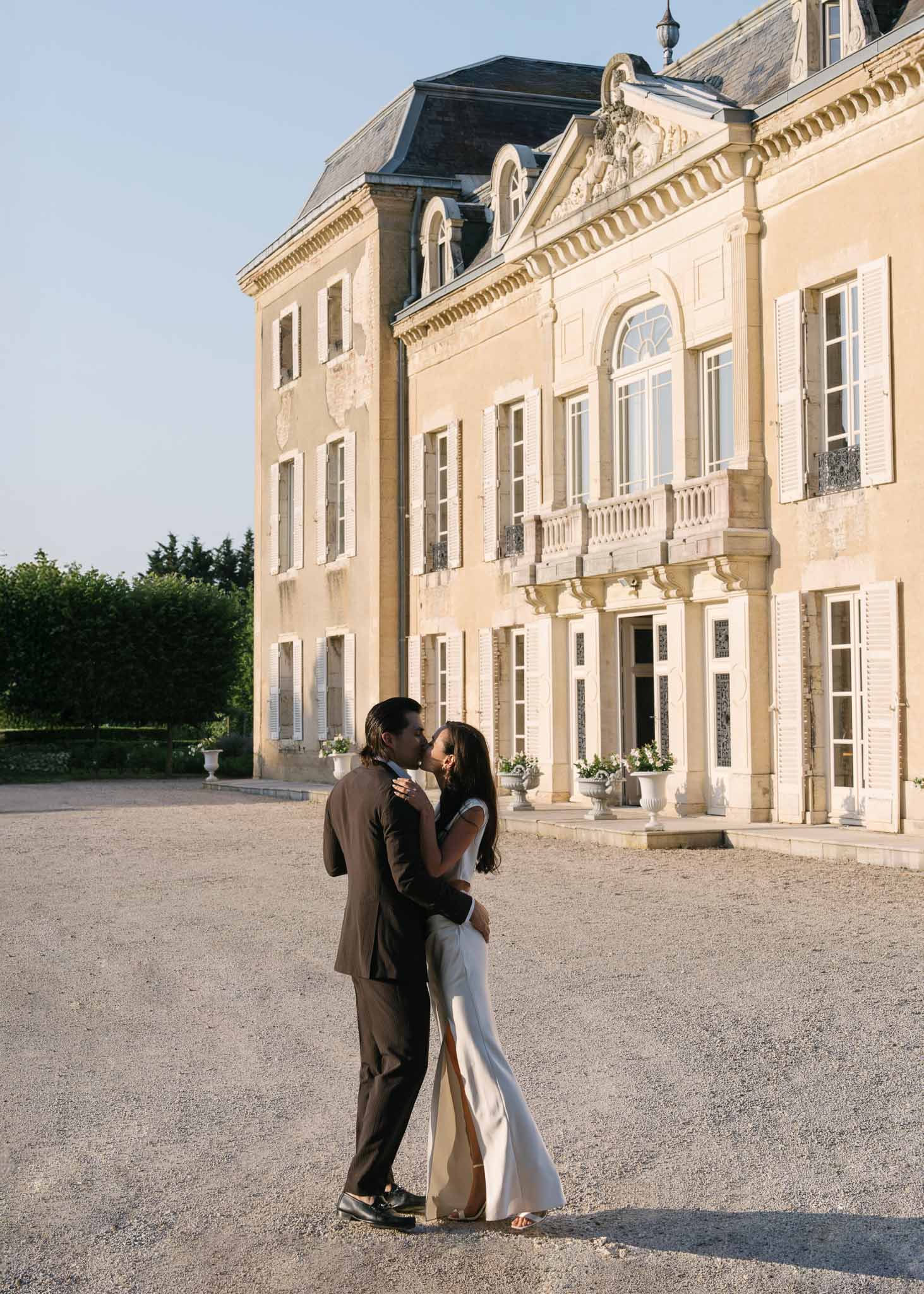 Couple kissing on chateau gravel forecourt at golden hour before cream stone facade with urn planters and balconies
