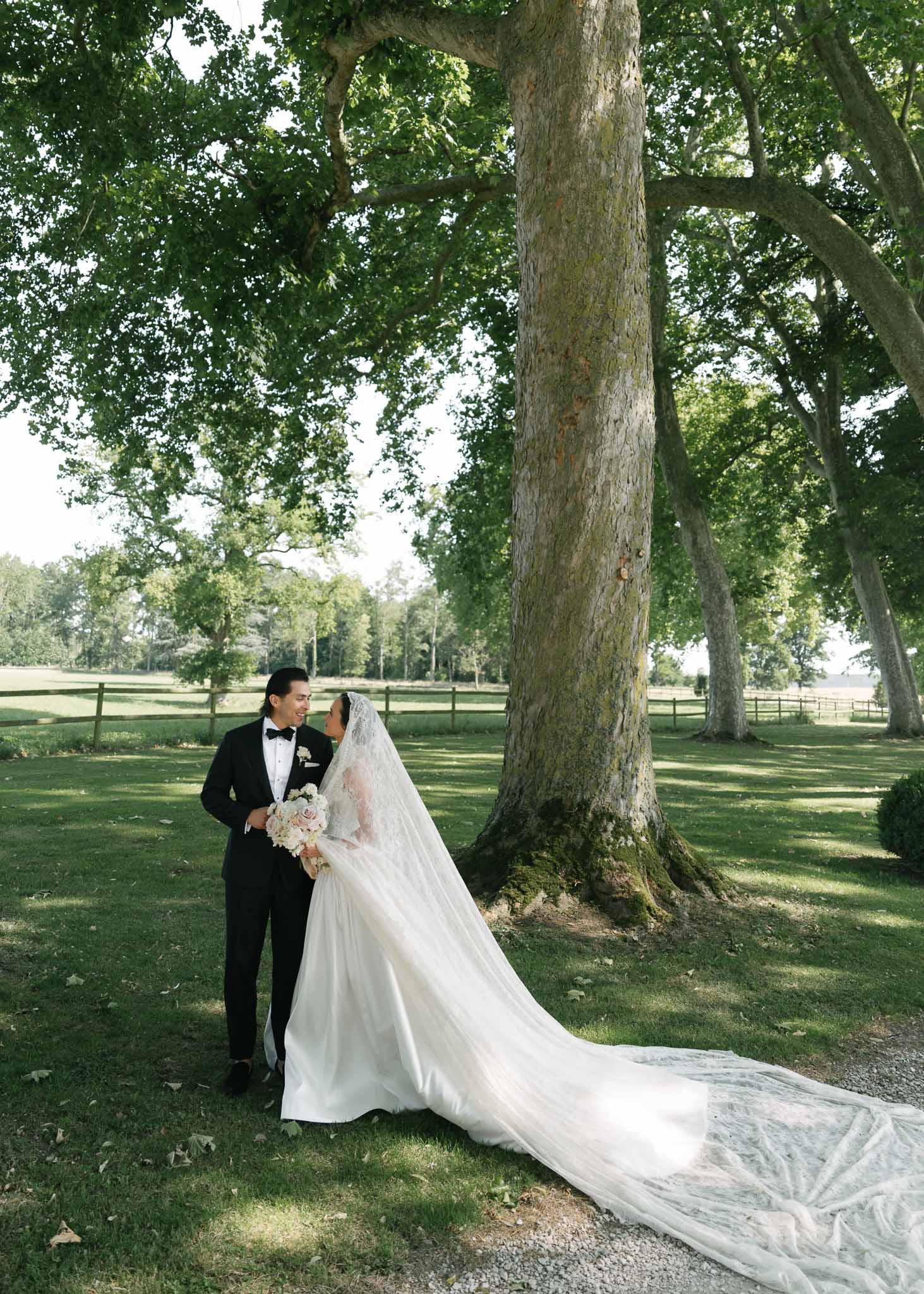 Bride in lace gown with ornate cathedral veil and groom in black tuxedo facing each other under plane tree