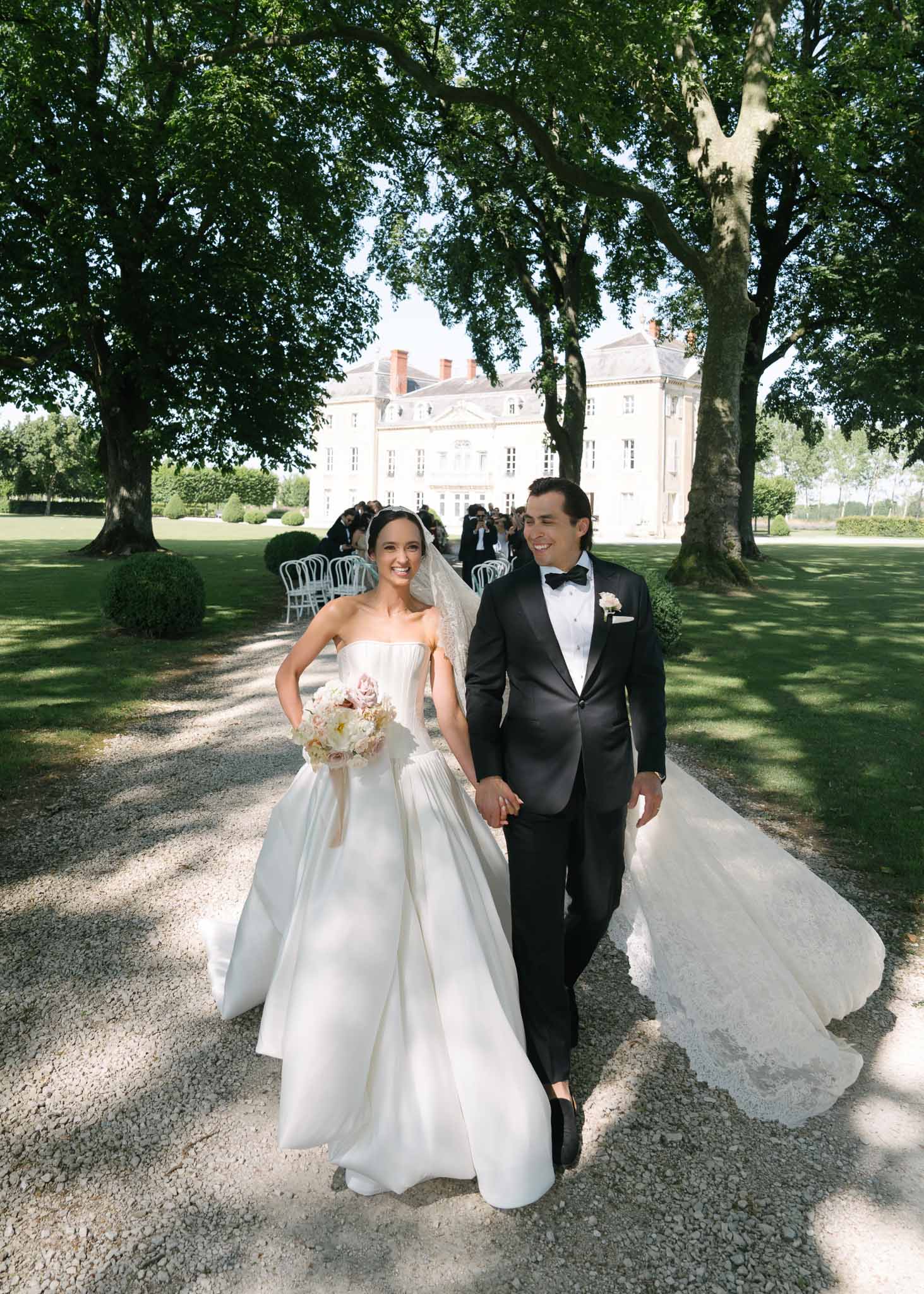 Couple walking from ceremony bride in ball gown with cathedral lace veil and blush peony bouquet