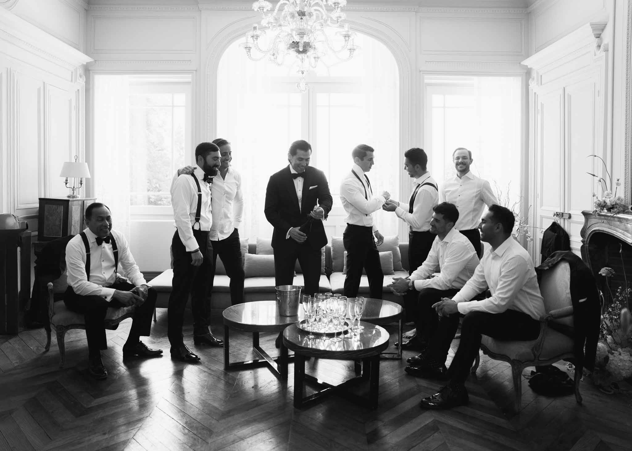 Groom and groomsmen in tuxedos gathering for champagne toast in chateau salon with chandelier