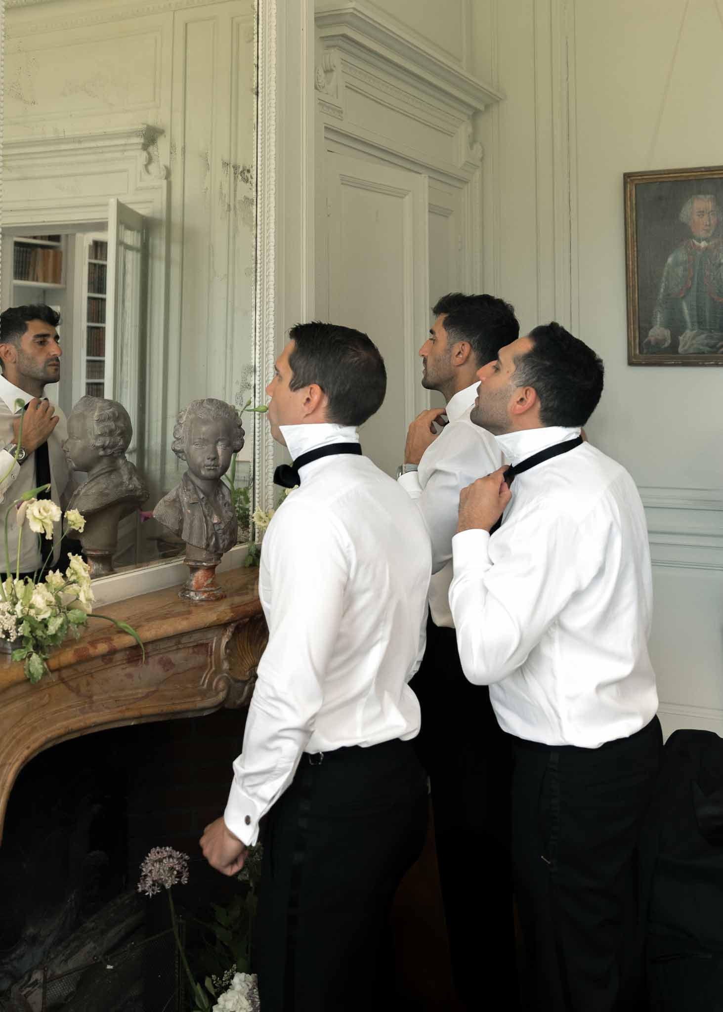 Three groomsmen adjusting black bow ties in front of ornate mirror in chateau room with marble fireplace