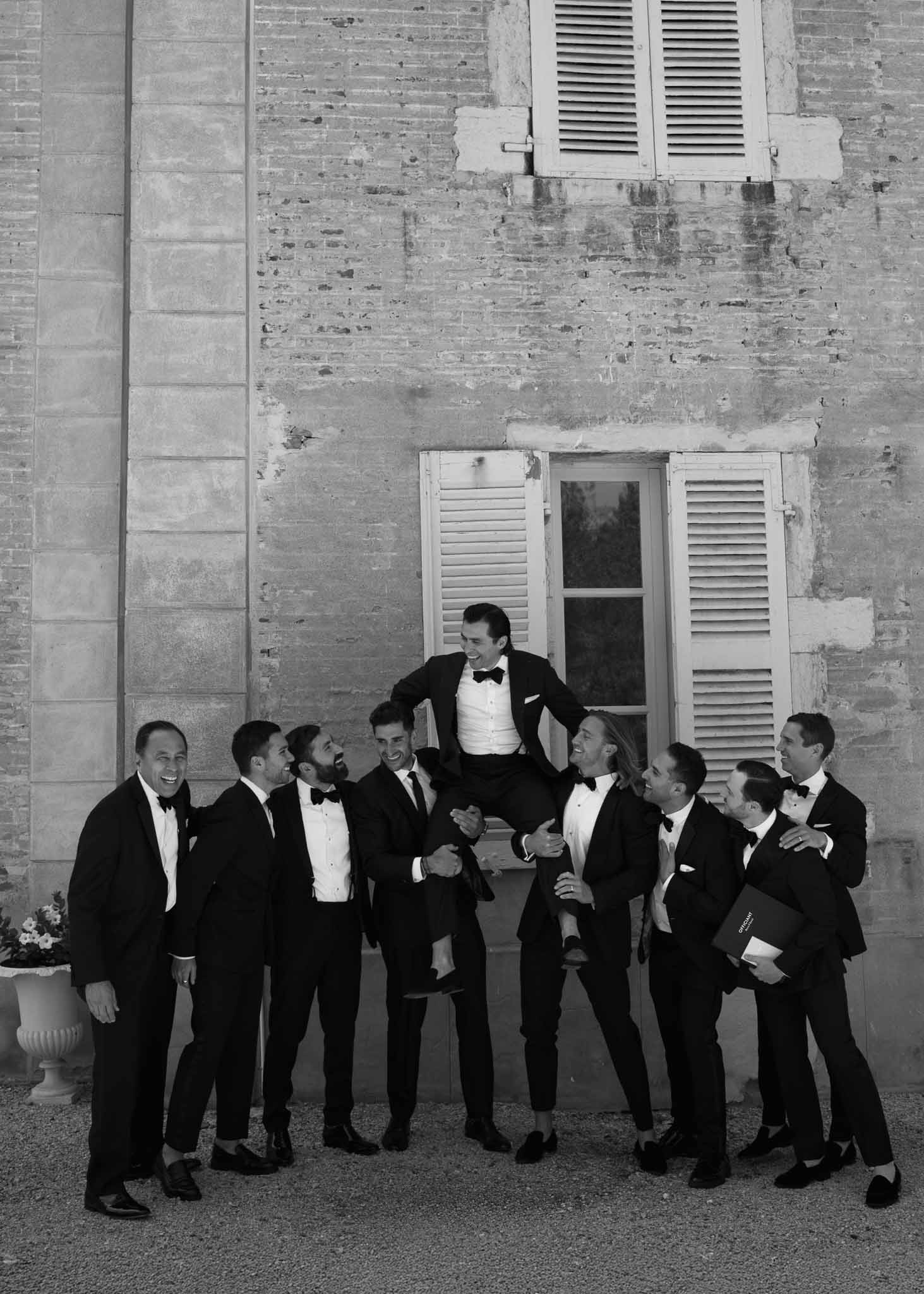 Nine groomsmen in tuxedos lift groom onto window ledge at chateau stone facade in B&W