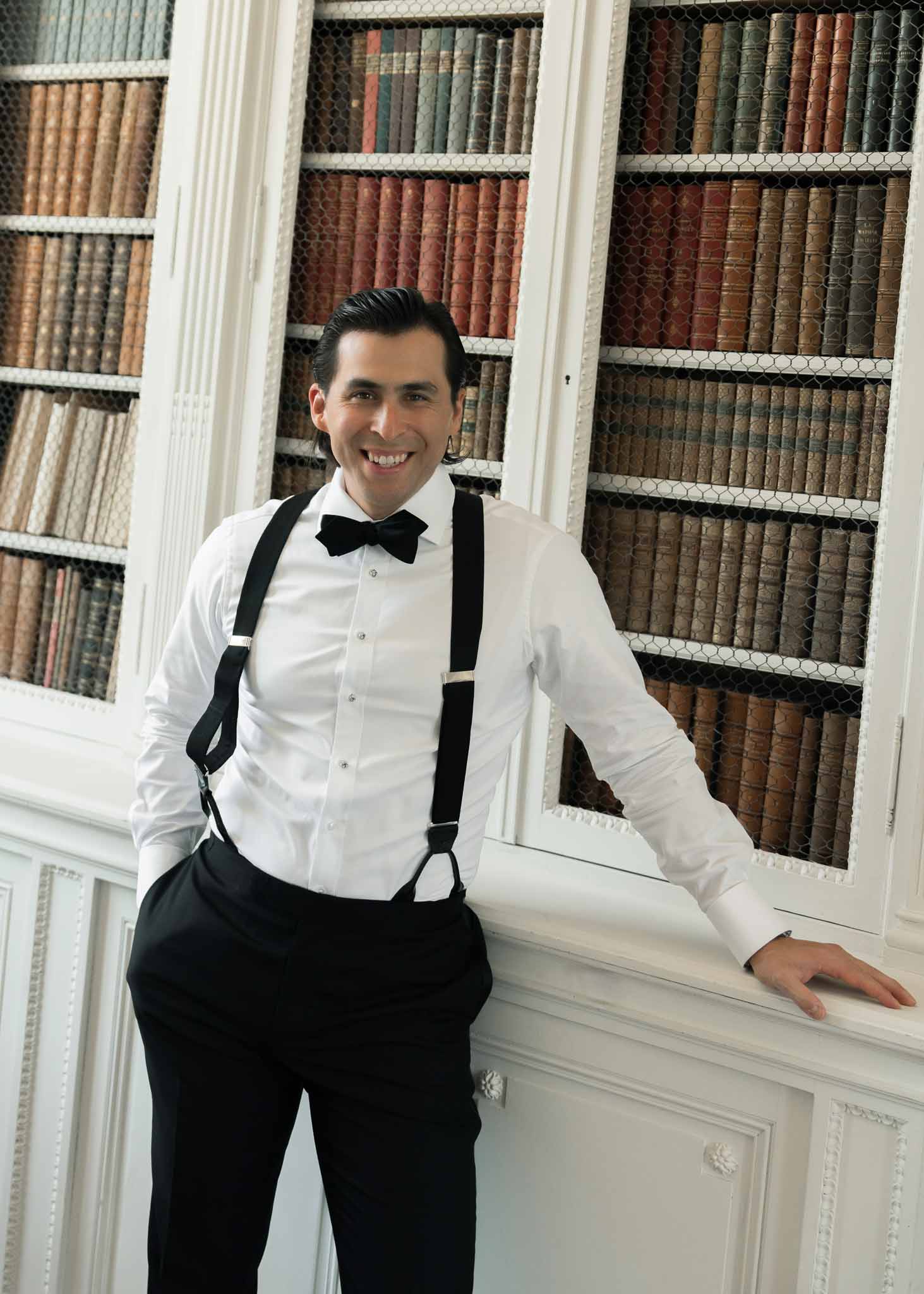 Groom in white shirt, black bow tie, and suspenders leaning against bookcase in chateau library