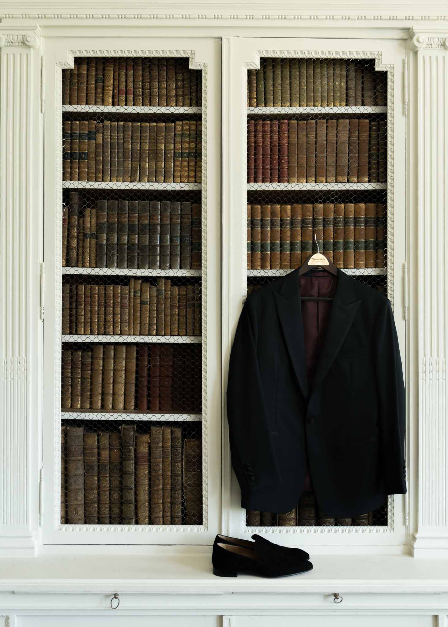 Navy suit jacket and velvet slippers hung on white bookcase with antique leather-bound books