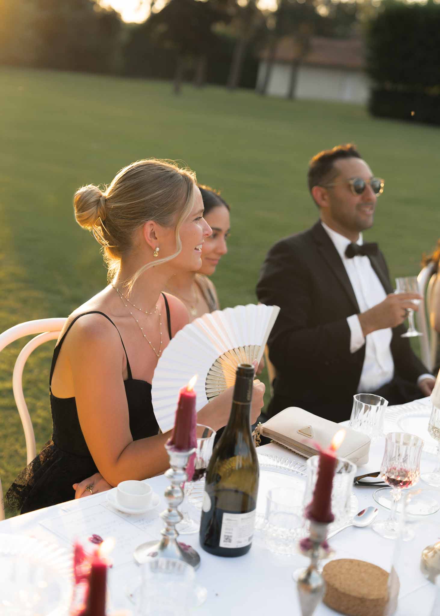 Reception guests at golden hour with burgundy taper candles, crystal glasses, and woman holding paper fan