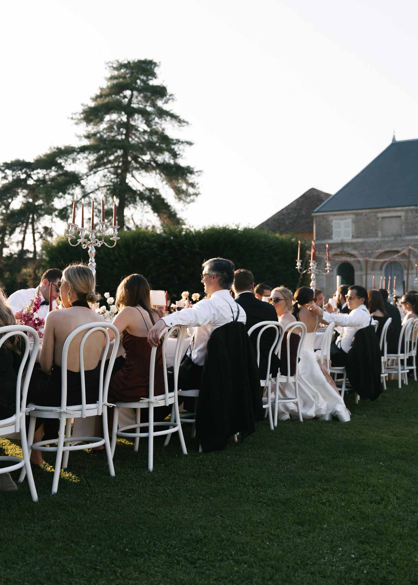 Twenty-five guests at long table with red taper candelabras and floral centerpieces before chateau at golden hour