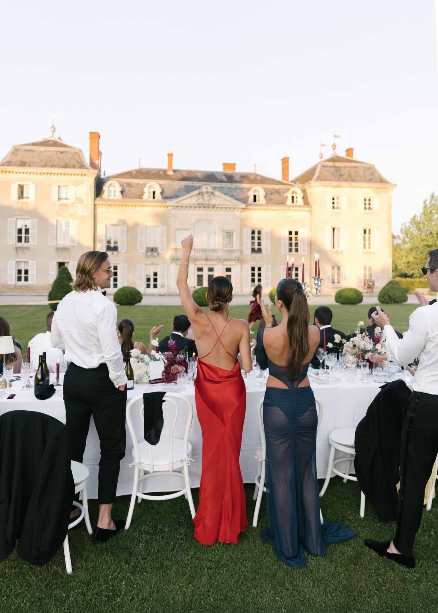 Golden hour reception with guests toasting at long tables with burgundy tapers and pink blooms before chateau