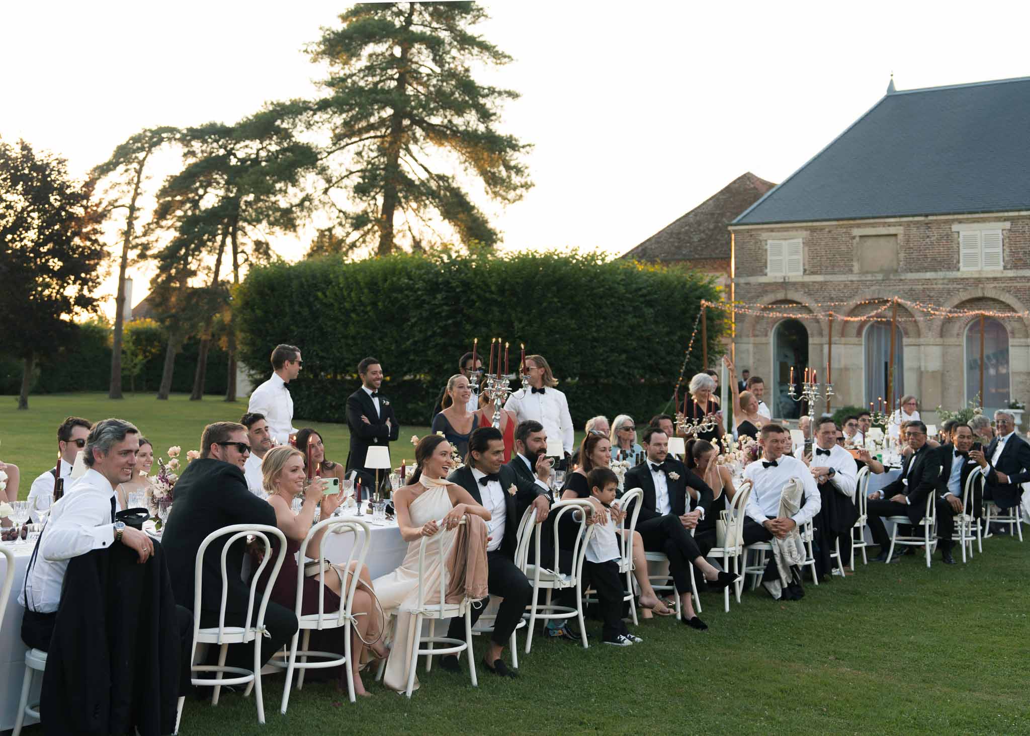 Black-tie guests at long tables with silver candelabras and fairy-lit stone arches at golden hour