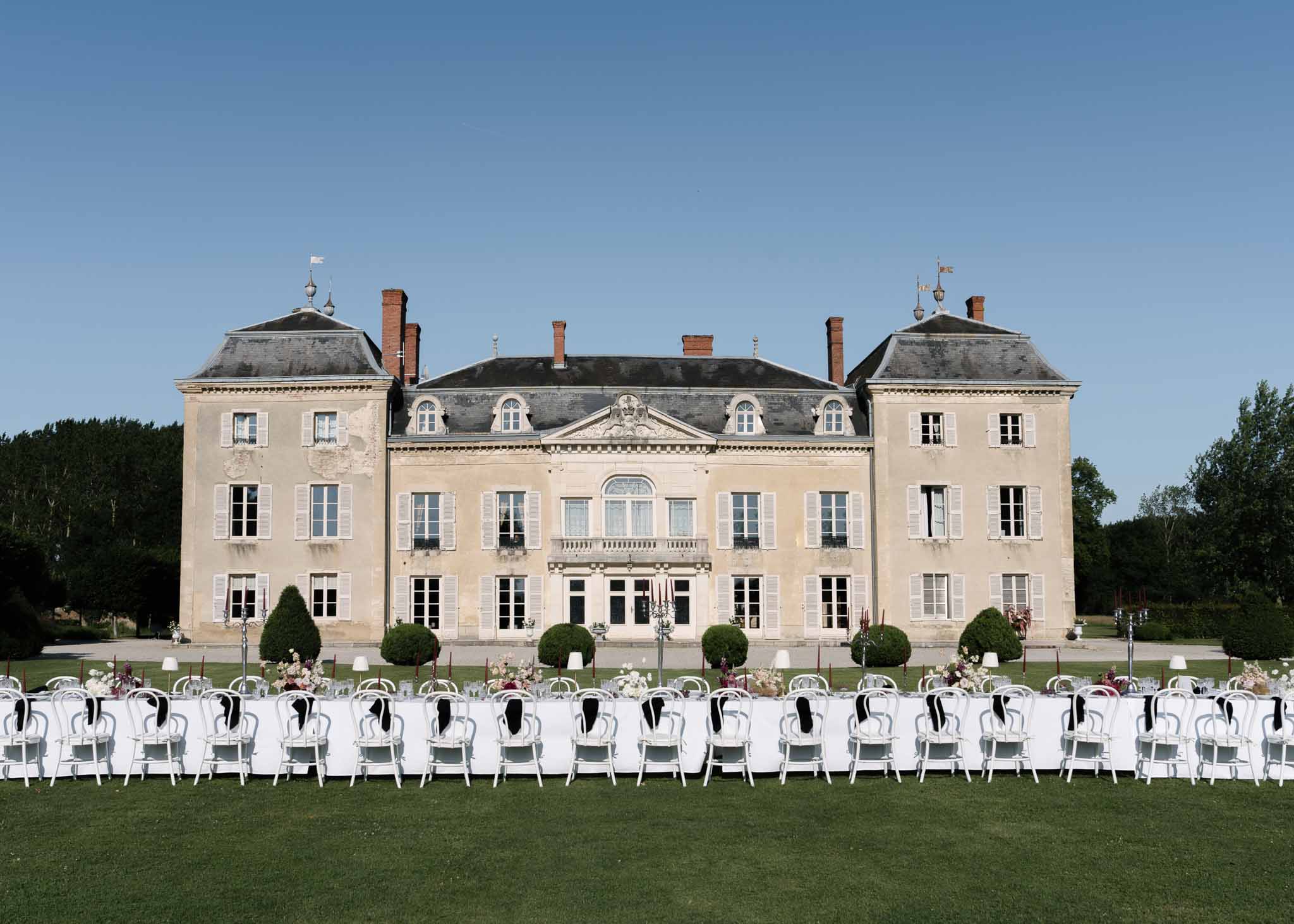 Long feasting table with white linen and candelabras on lawn in front of French chateau facade