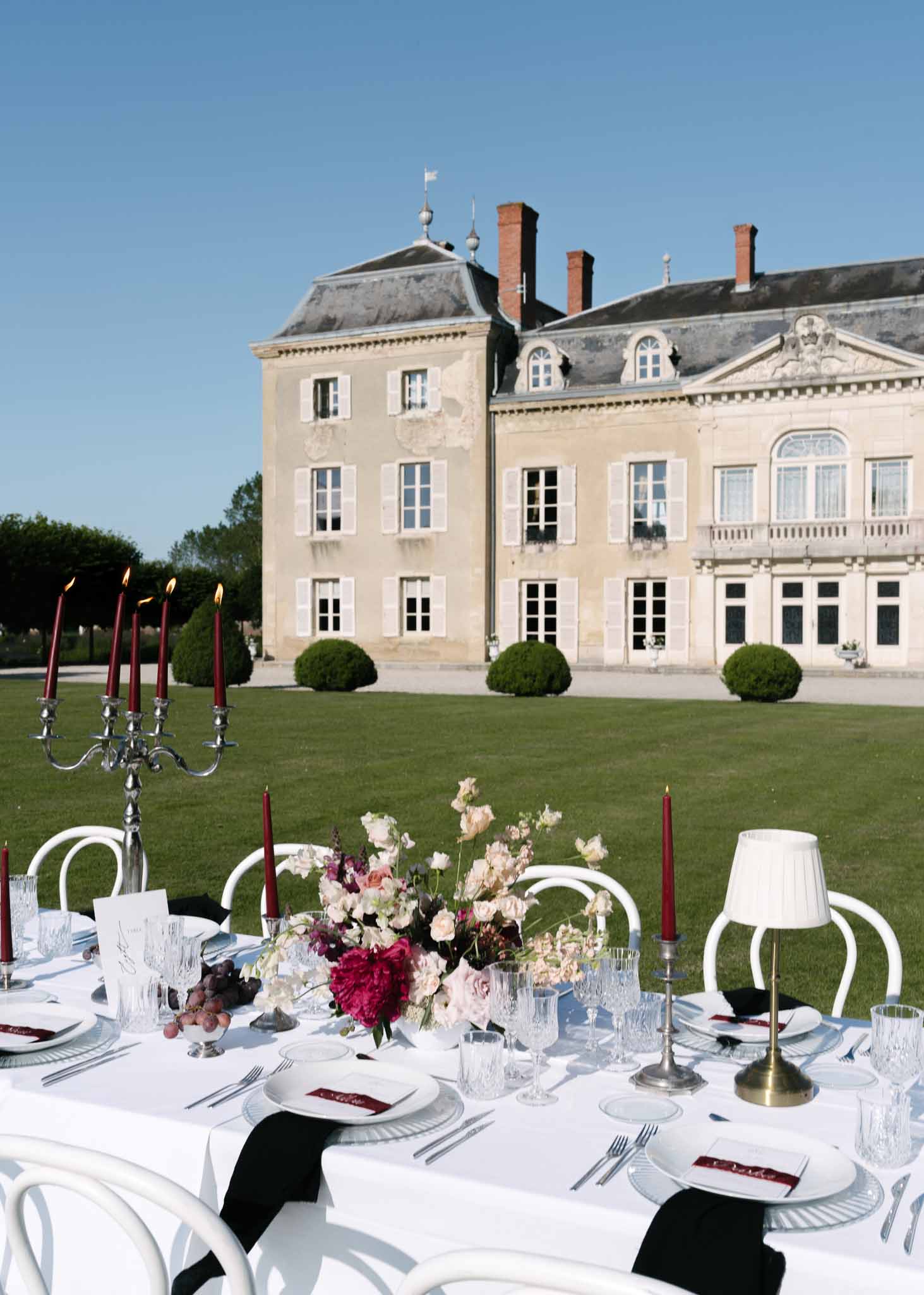 Outdoor reception table with magenta peonies, burgundy candles, silver candelabra before French chateau