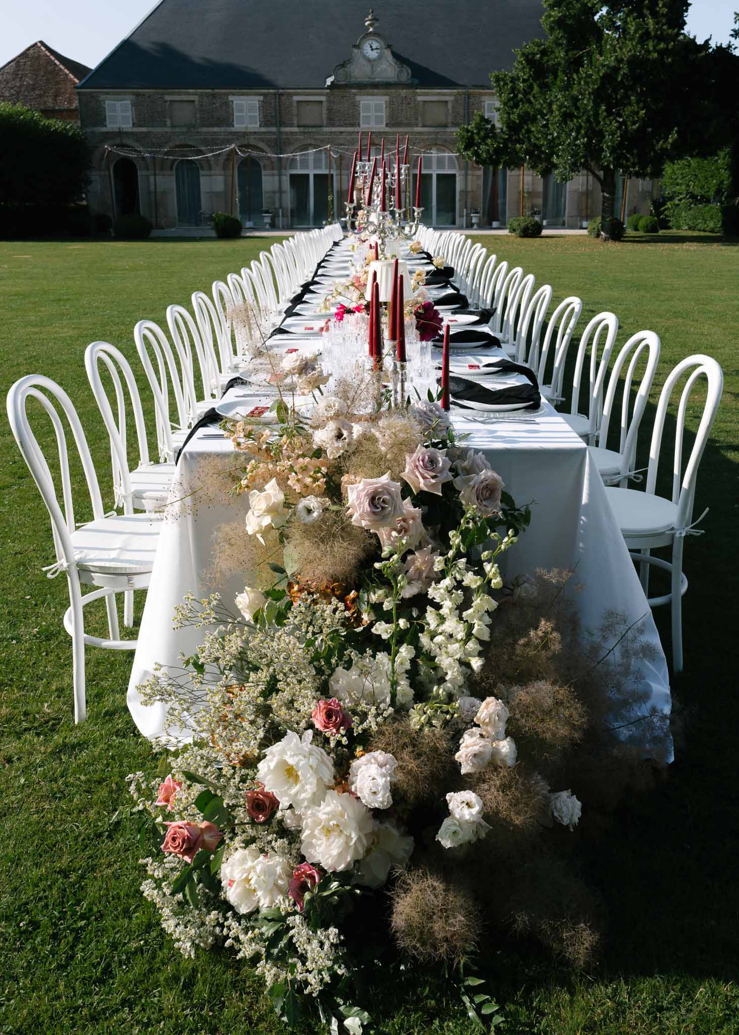 Long table with peony and rose runner, burgundy tapers, and black napkins before clock-pediment manor