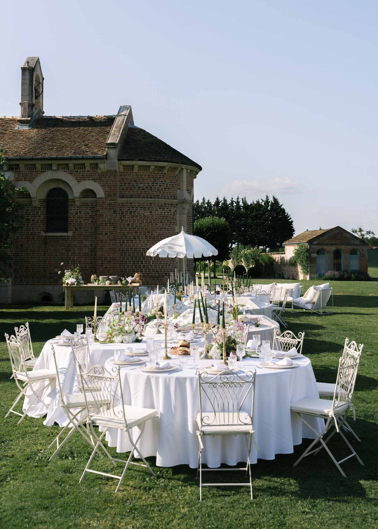 Round tables with brass candlesticks, green tapers, and white hydrangeas before French brick chapel