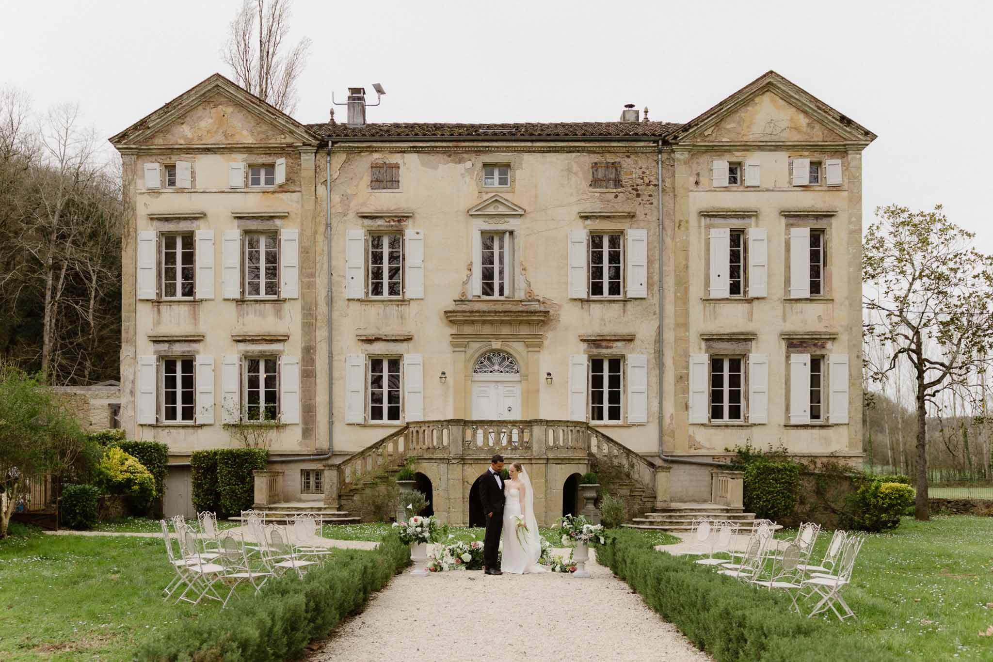 Bride and groom at base of chateau double staircase with ceremony chairs and white floral urns on gravel path