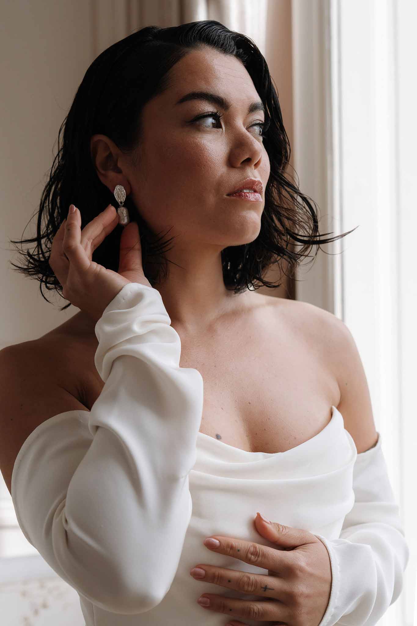 Bride adjusting silver drop earring by a window in an off-the-shoulder white gown with balloon sleeves