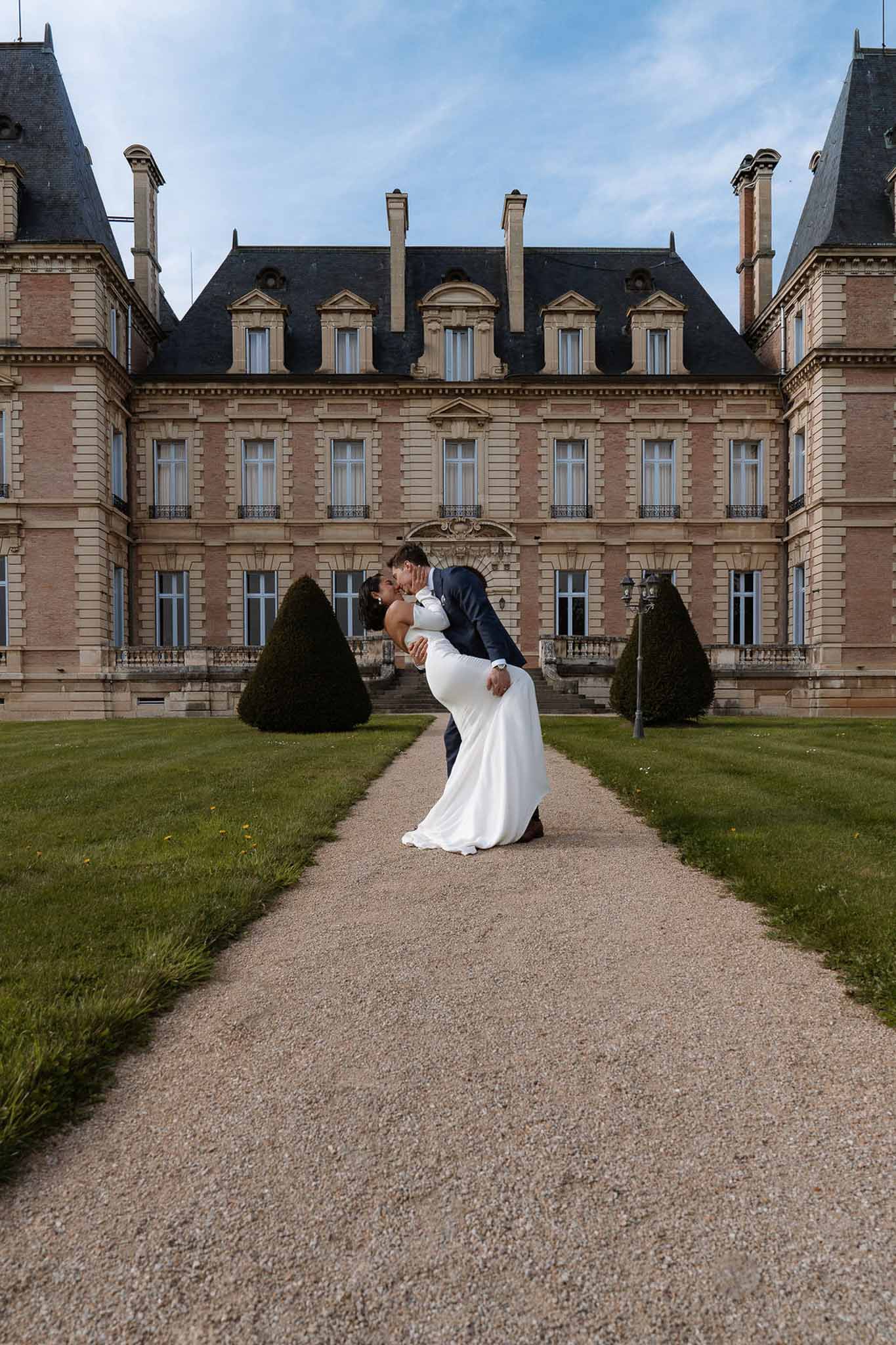 Groom in navy suit dipping bride for a kiss on gravel pathway with pink-brick French chateau in background