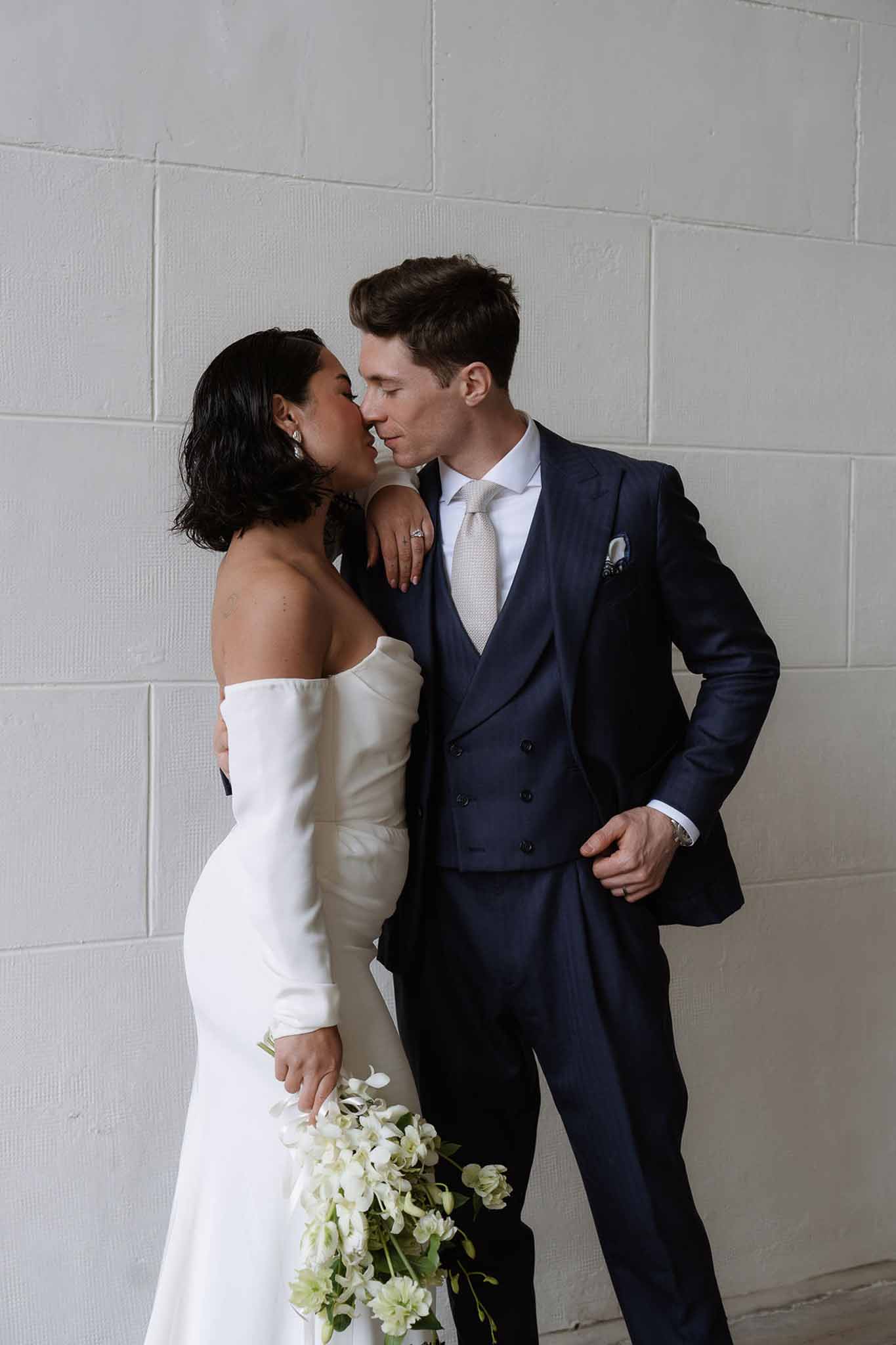 Bride in off-shoulder white gown kissing groom in navy three-piece suit holding sweet pea bouquet against white wall