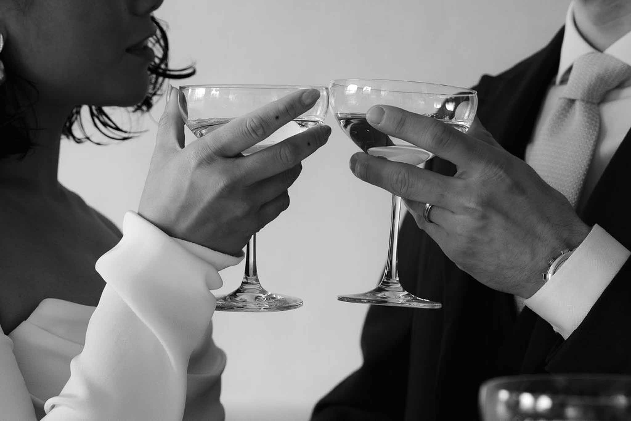 Black-and-white close-up of bride and groom toasting with coupe champagne glasses showing hands and rings
