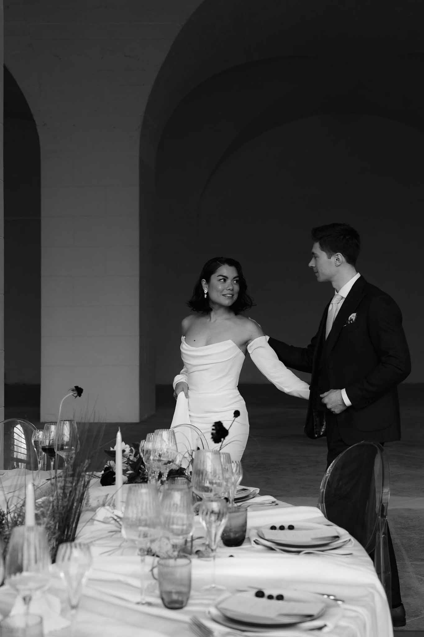 Black and white couple at head table with taper candles and dark blooms in vaulted stone venue