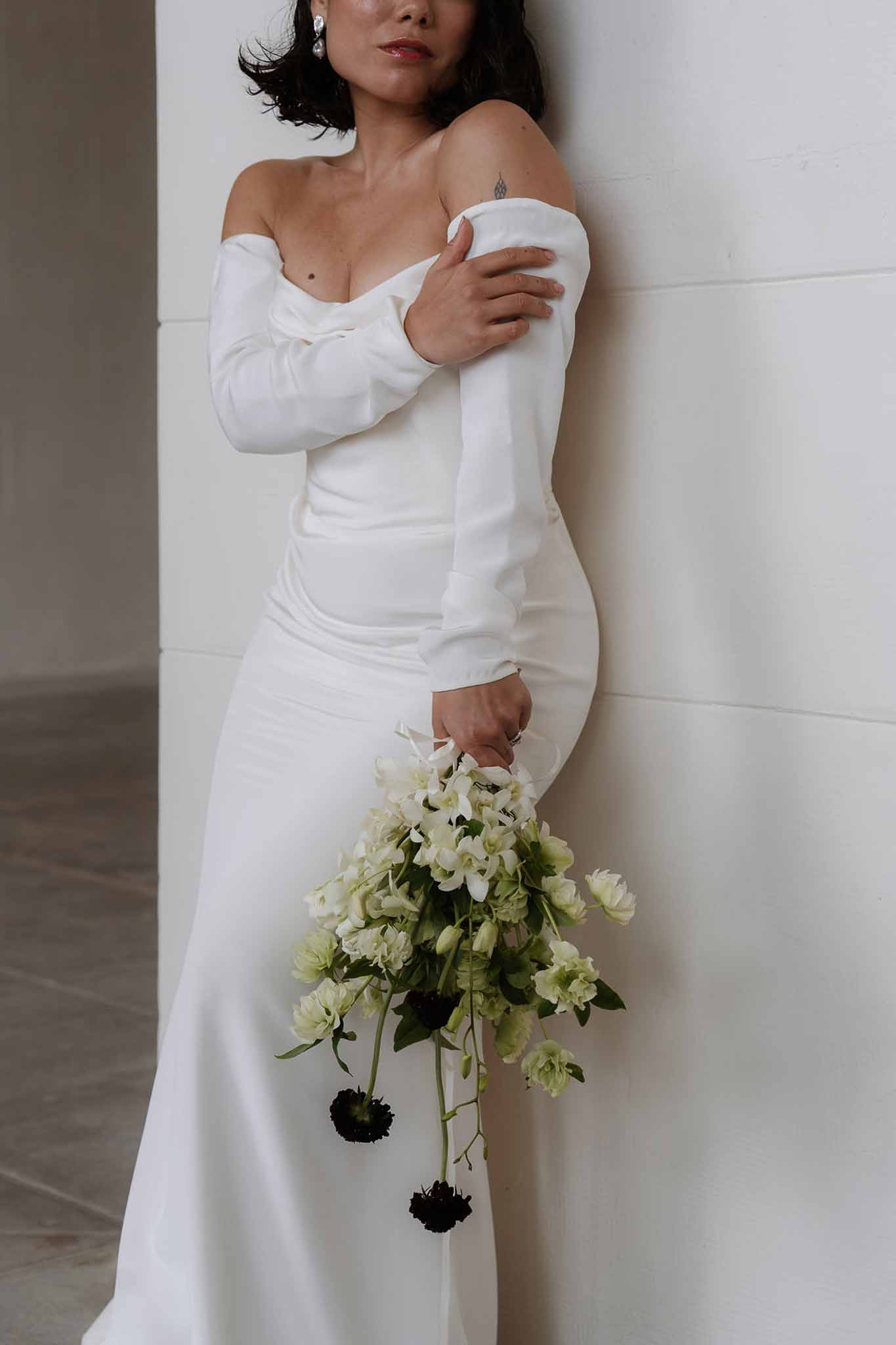 Bride in off-shoulder white gown holding trailing orchid, ranunculus, and dark scabiosa bouquet against white wall