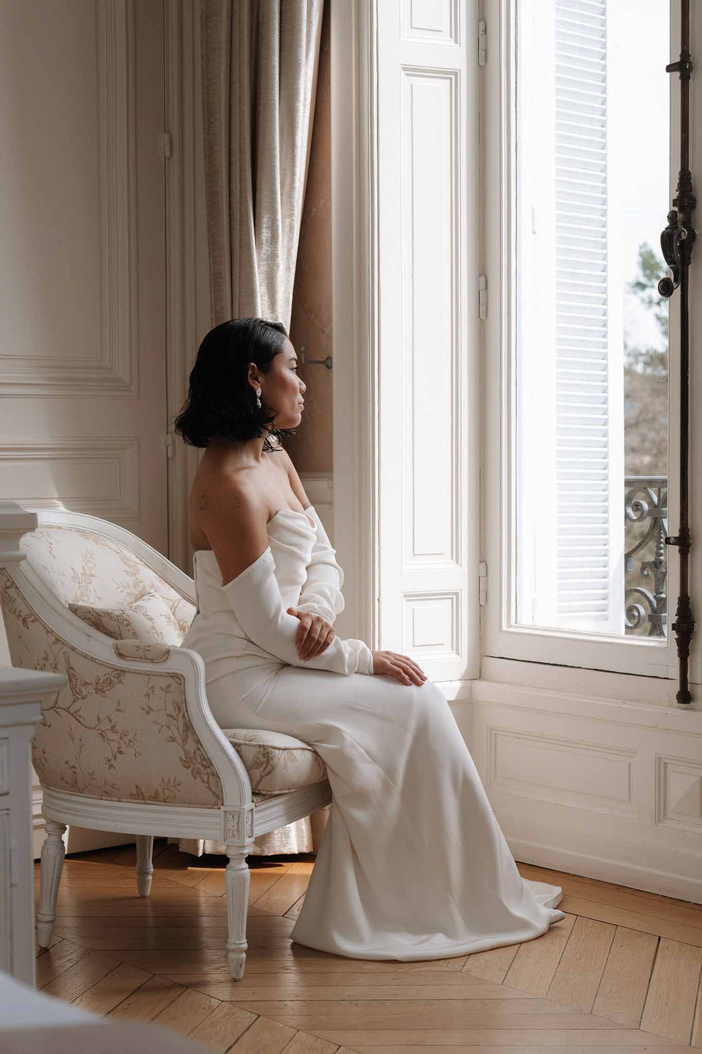 Bride in off-shoulder gown with bob haircut seated in botanical armchair by louvred window in salon