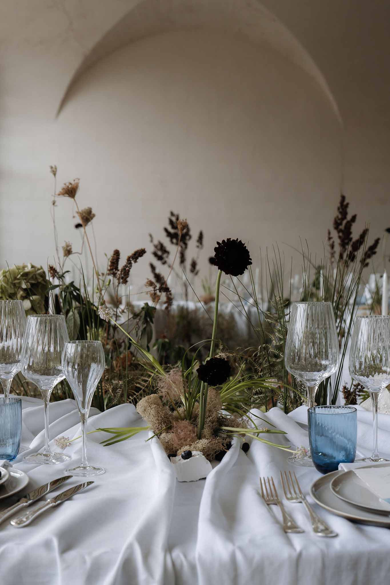 Place setting with gold flatware, dusty blue glasses, and burgundy scabiosa botanical centerpiece under vaulted arches