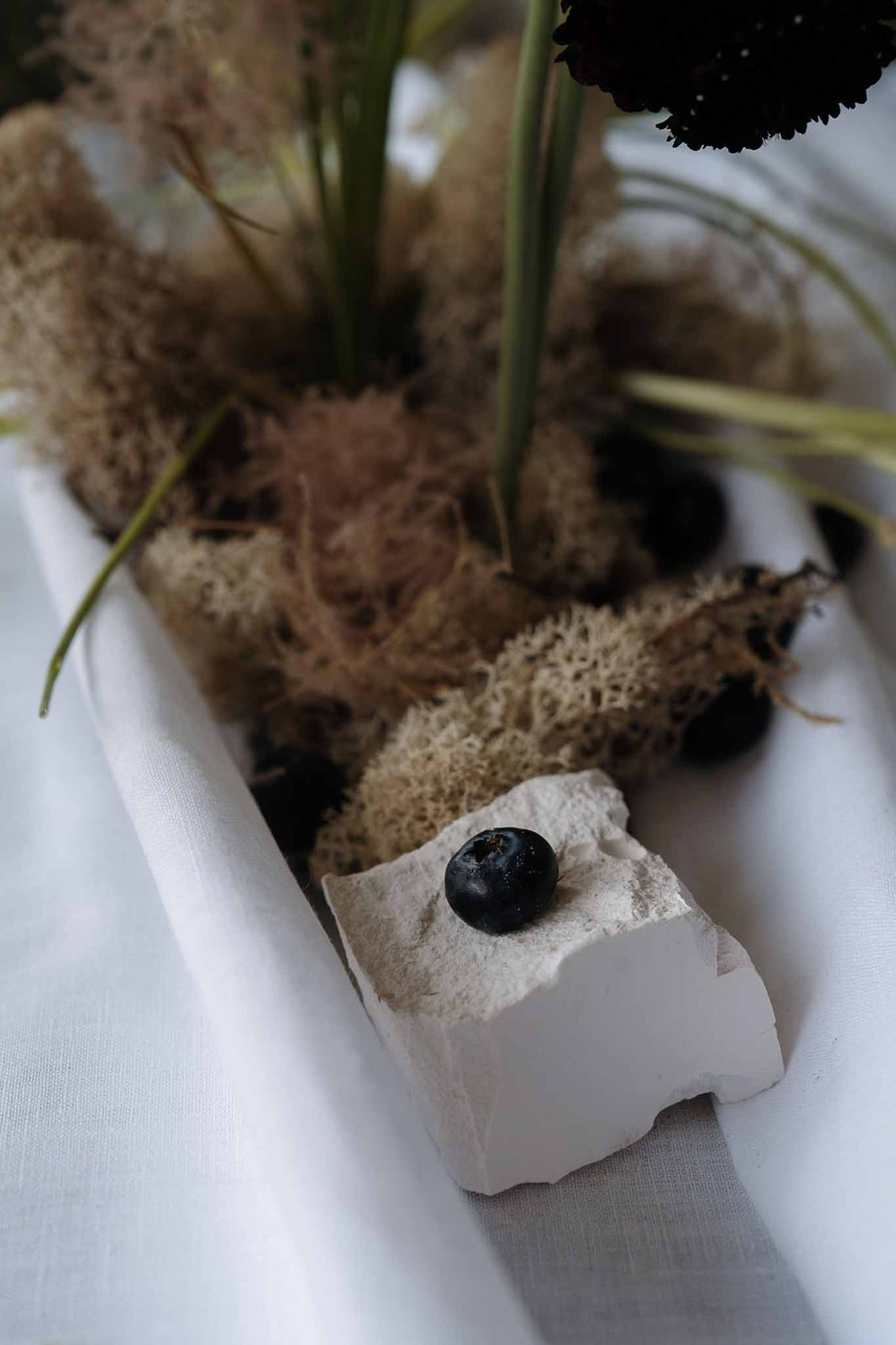 Moody table centerpiece with dried moss, dark stones, green stems, and burgundy scabiosa flower