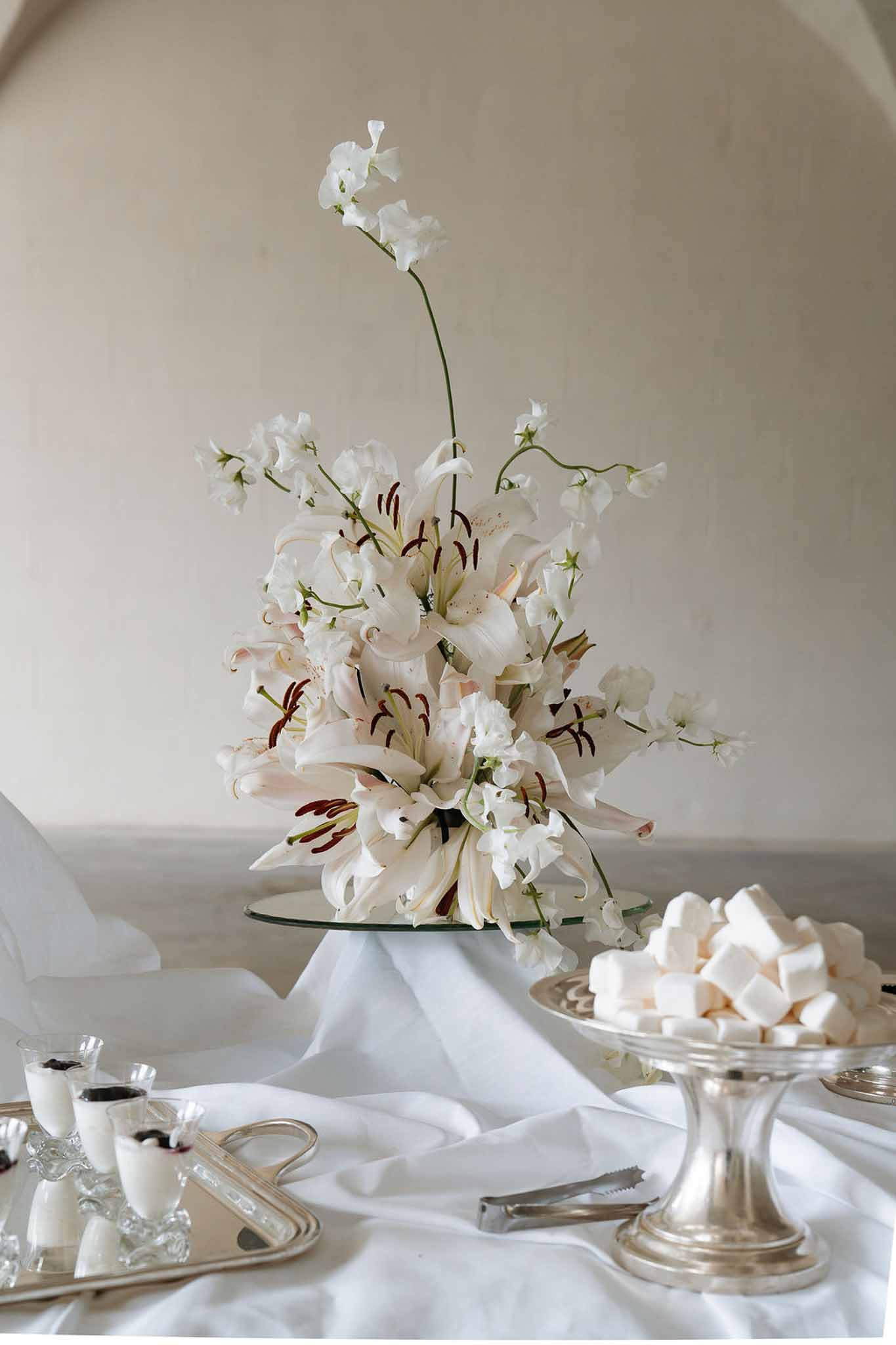 Dessert table with white lilies on glass stand, pink meringues on silver pedestal, and berry-topped cream shots