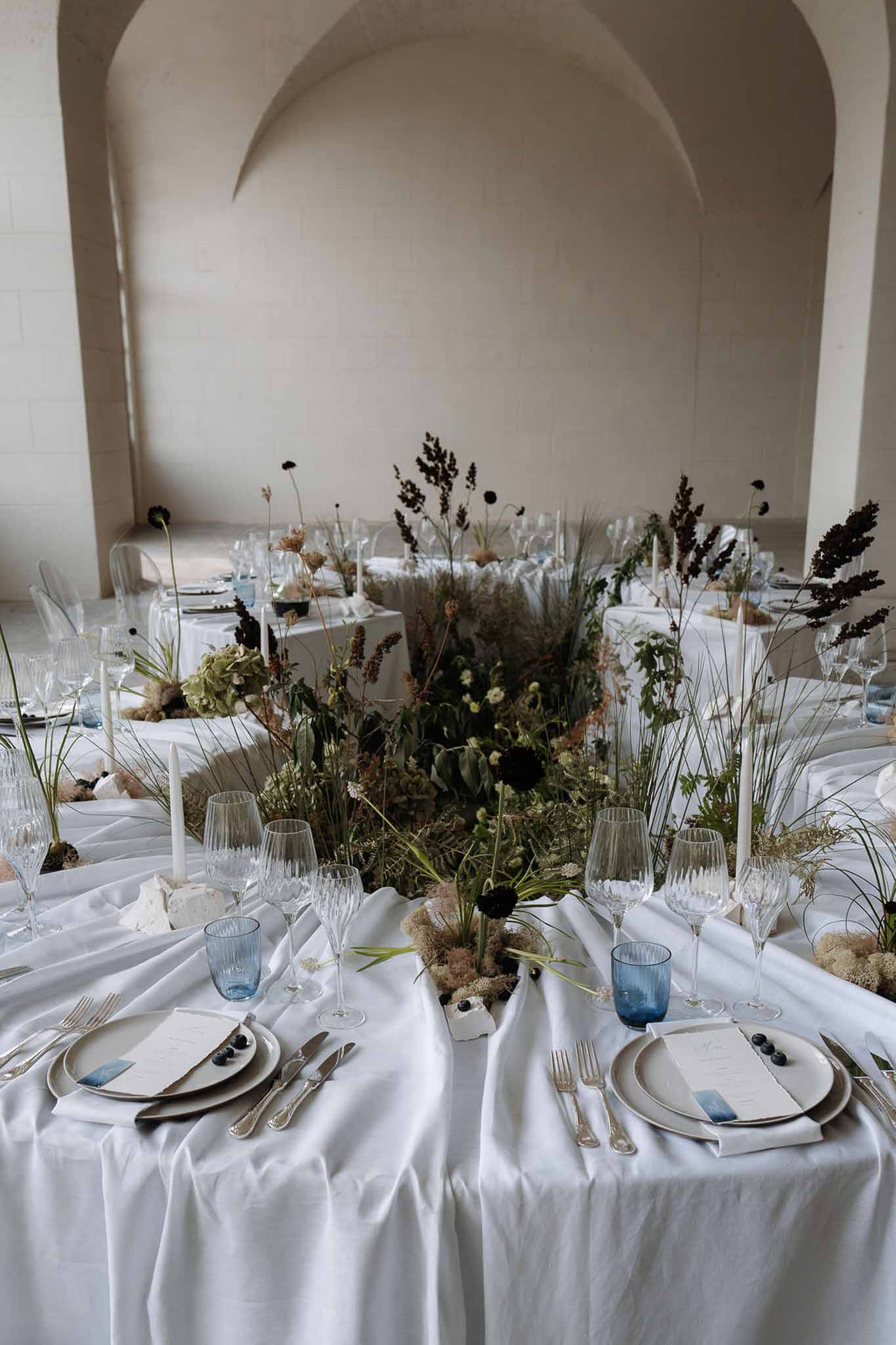 Reception tablescape with taupe plates, dusty blue glasses, burgundy wildflowers, ferns, and white taper candles under sto...
