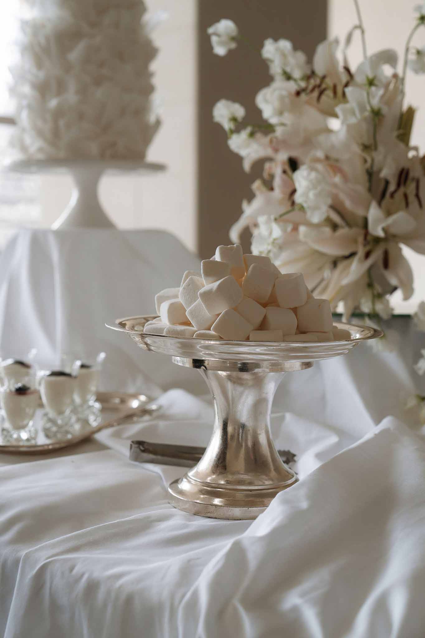 Close-up of a silver pedestal holding marshmallows beside a ruffled white wedding cake and white lily floral arrangement