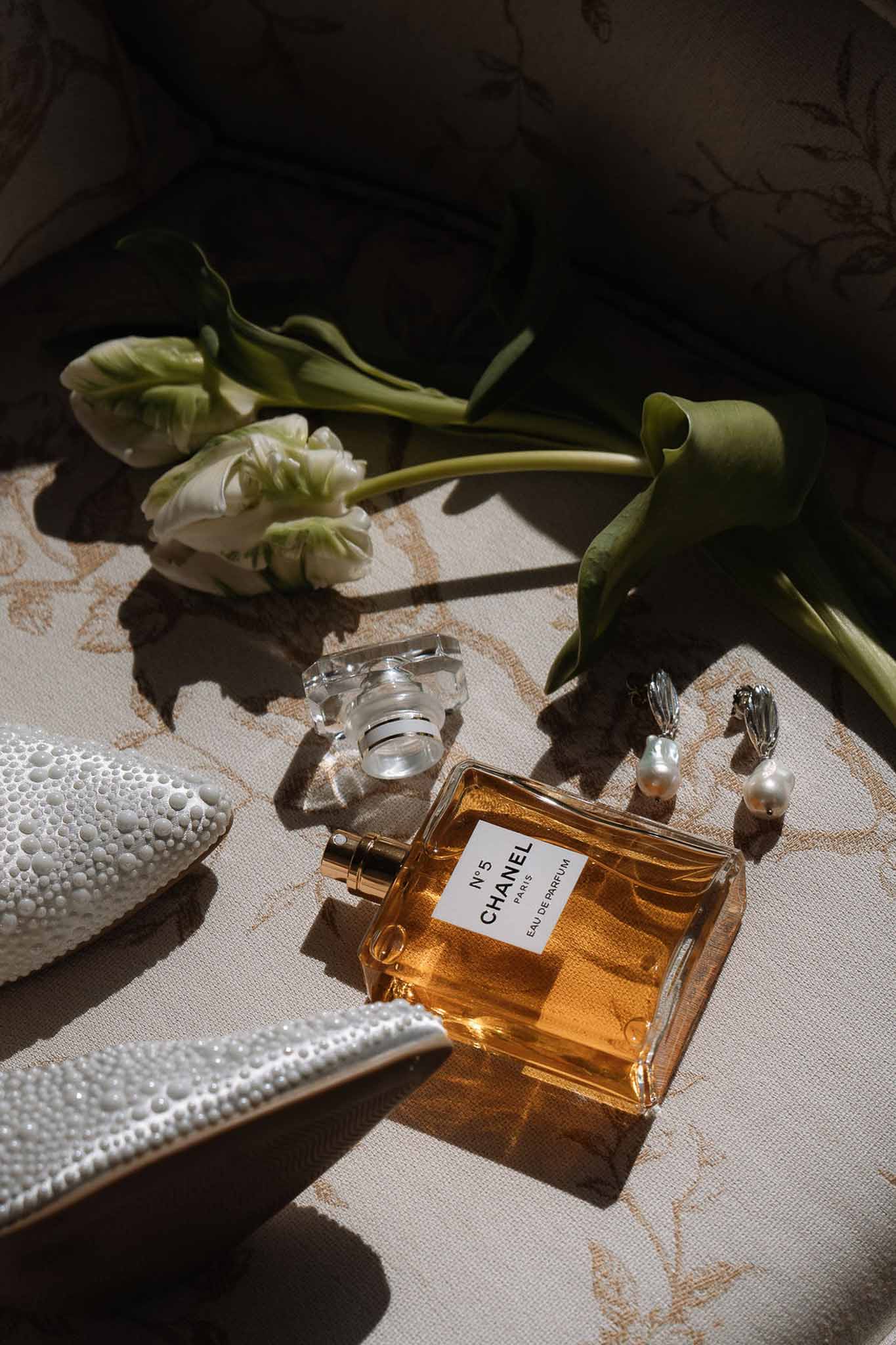 Flat lay of Chanel No. 5, baroque pearl earrings, white beaded clutch, and parrot tulips