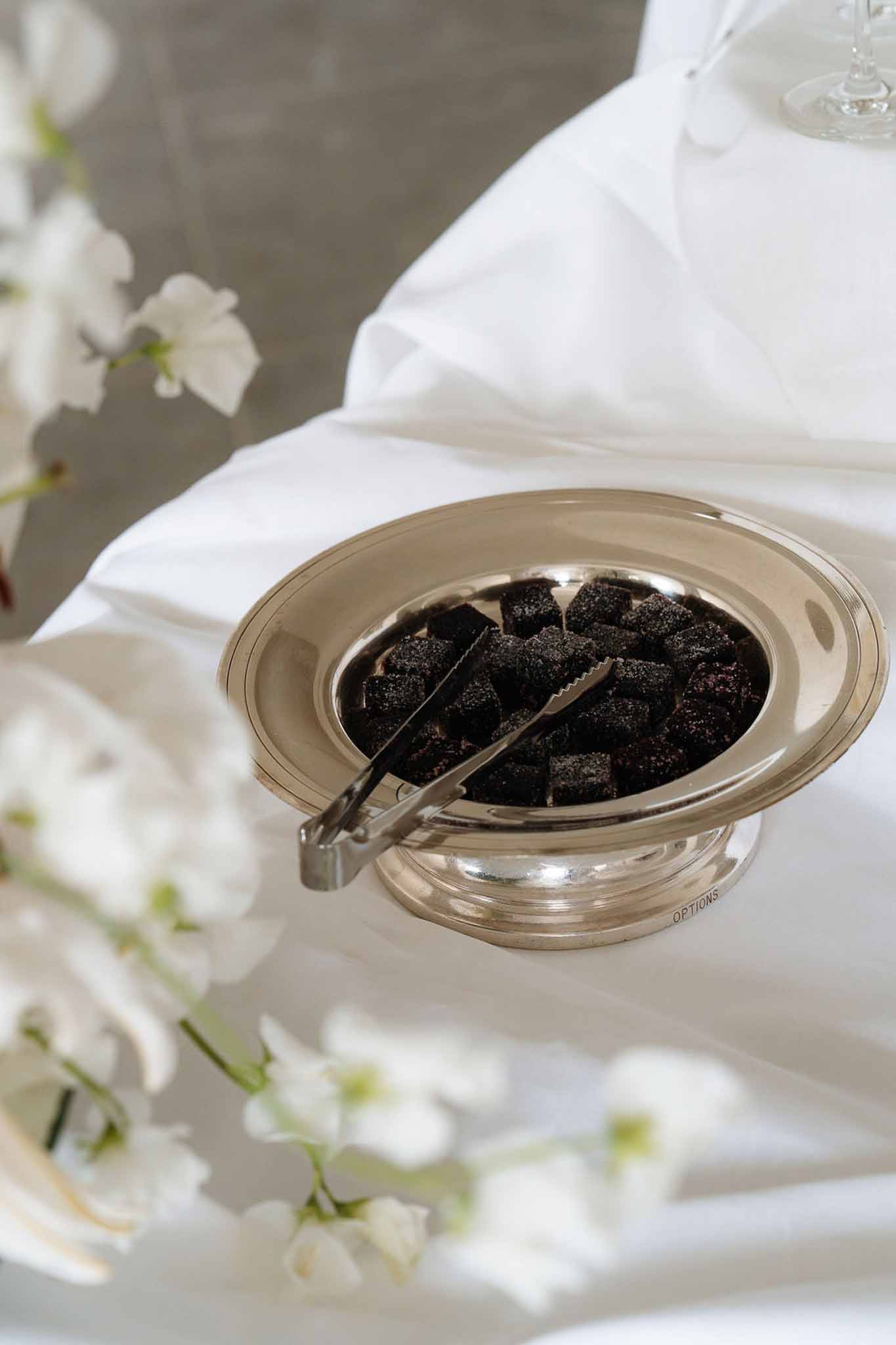 Silver pedestal bowl filled with dark chocolate confections with serving tongs on white linen beside orchids
