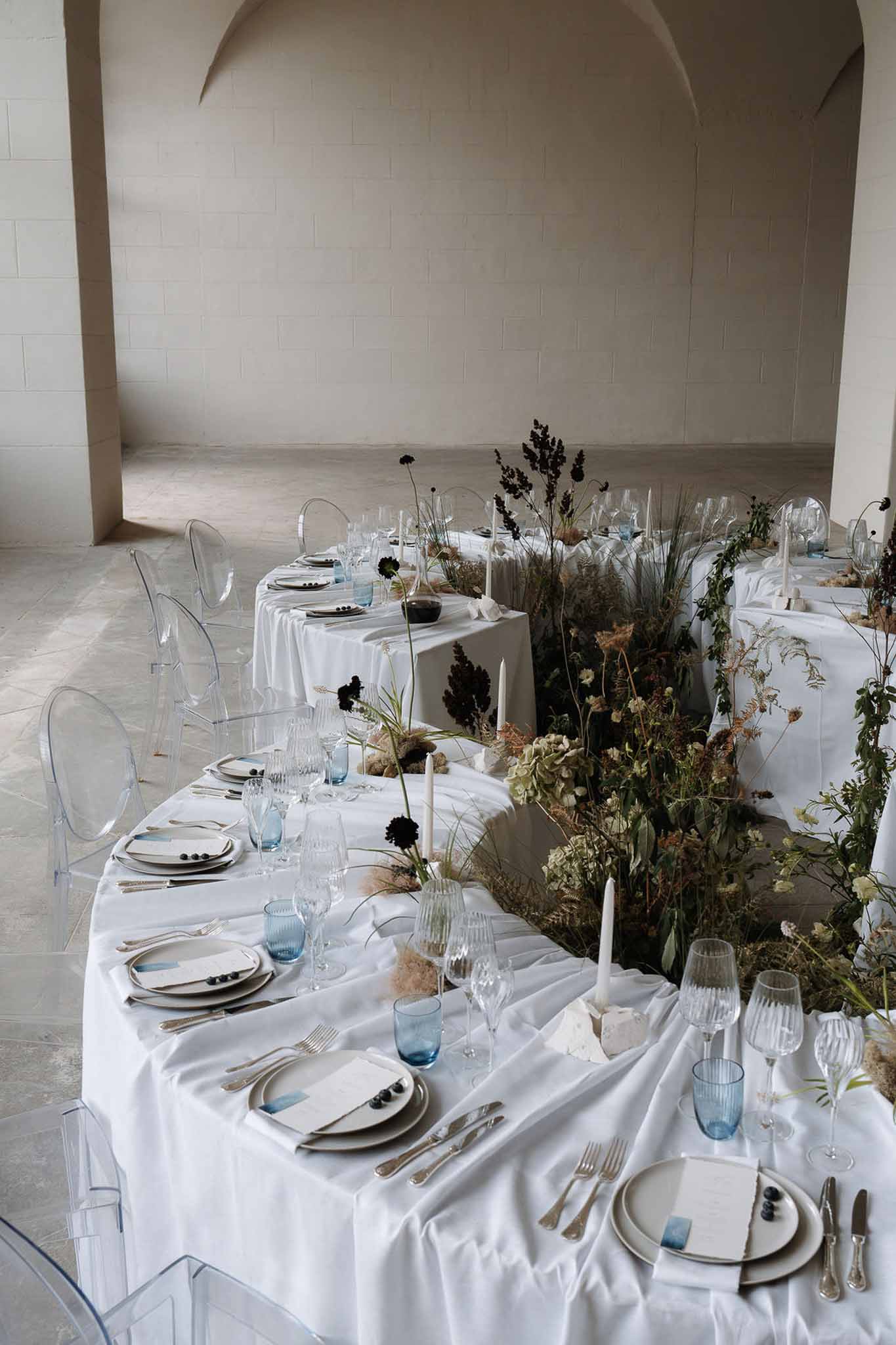 Minimalist reception tables with ghost chairs, pampas grass, burgundy scabiosa, and blue glassware in a stone venue