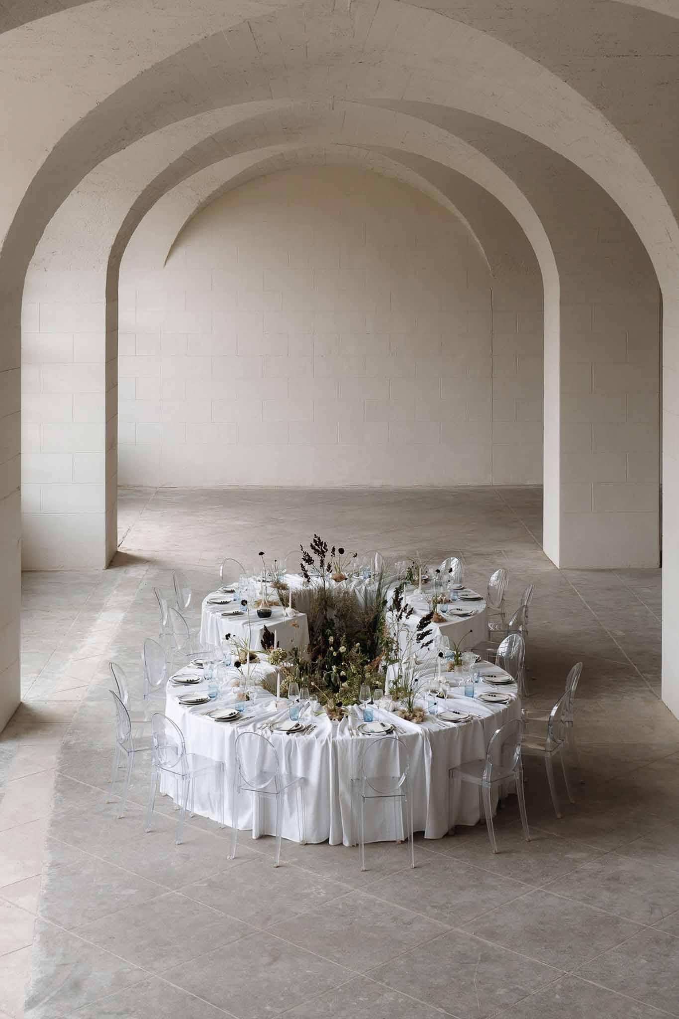 Vaulted stone hall reception with ghost chairs white linen tall dried grasses and meadow-style botanical centerpieces