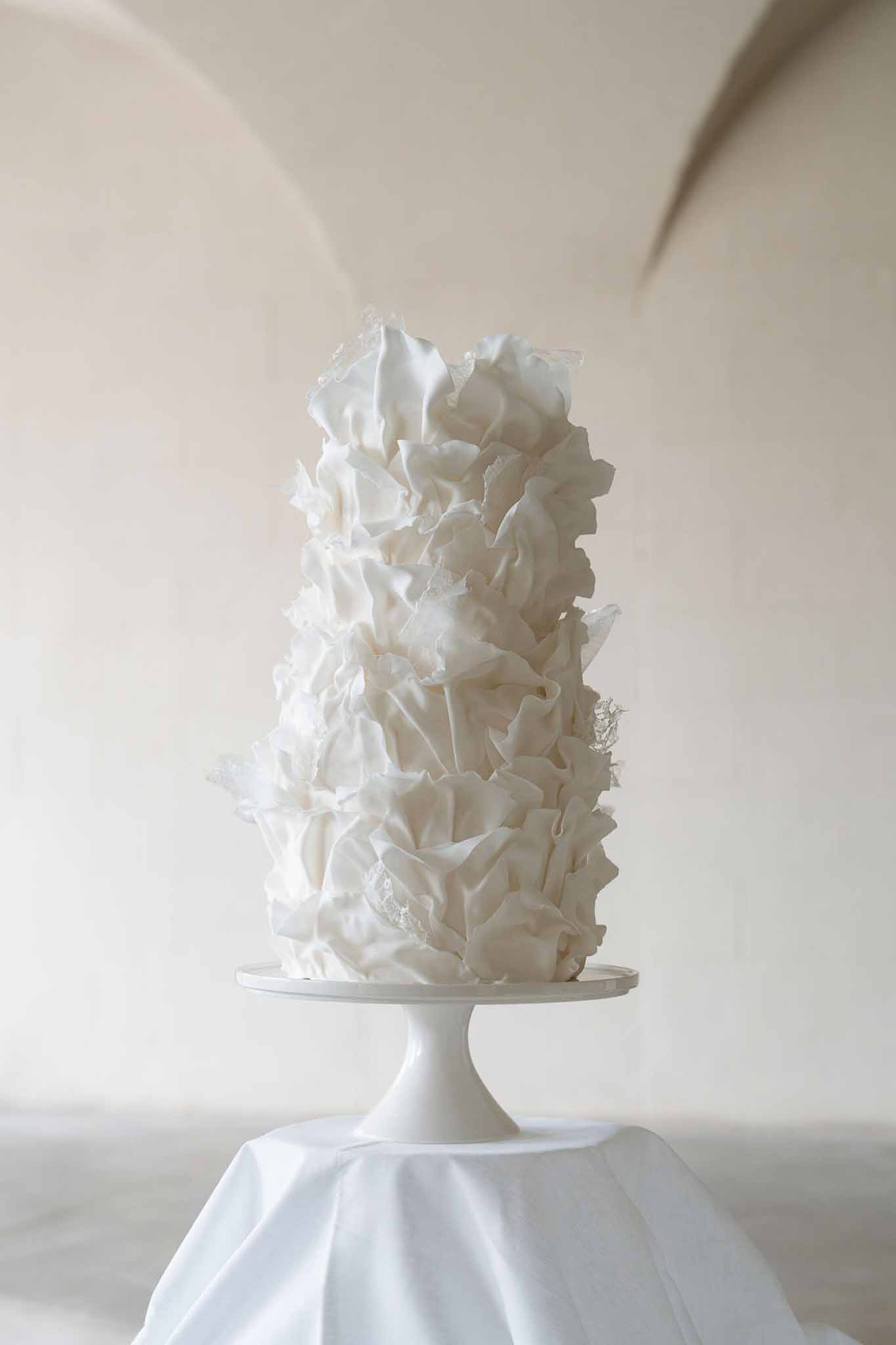All-white ruffled petal wedding cake on white pedestal stand with translucent sugar accents in arched indoor venue