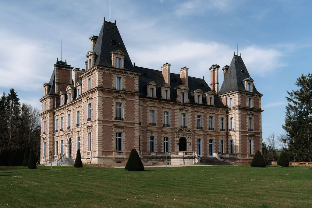 Pink-brick French chateau with slate mansard roofs, corner towers, dual staircases, and conical topiary on manicured lawn