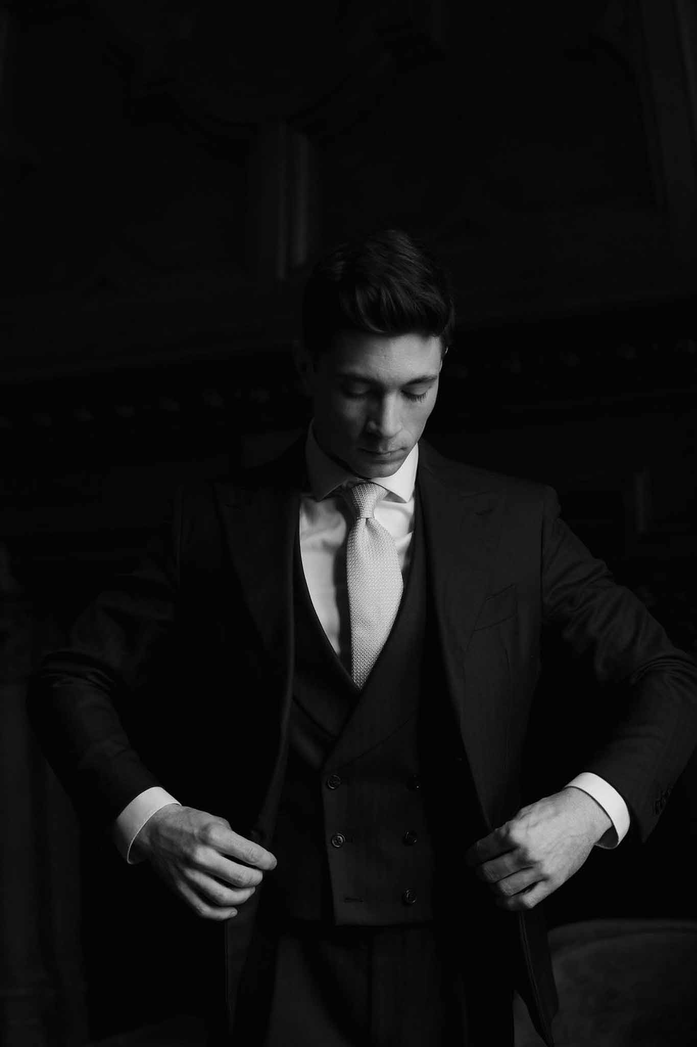 Black-and-white portrait of groom adjusting suit jacket with dramatic chiaroscuro lighting
