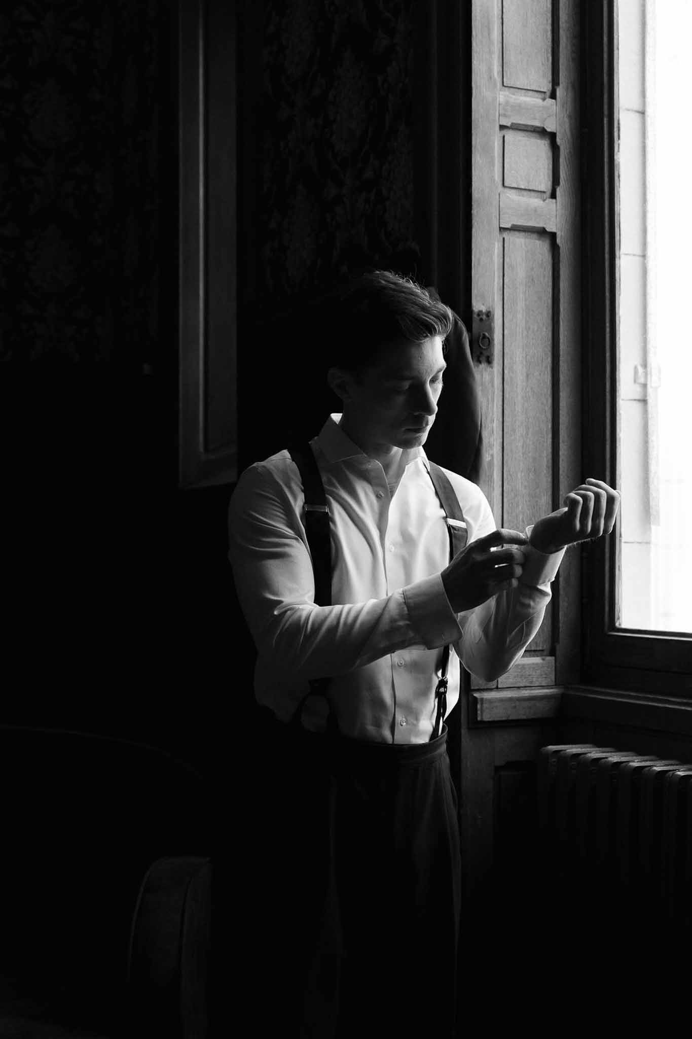 Groom fastening cufflinks by a tall shuttered window in dramatic black-and-white light at a historic venue