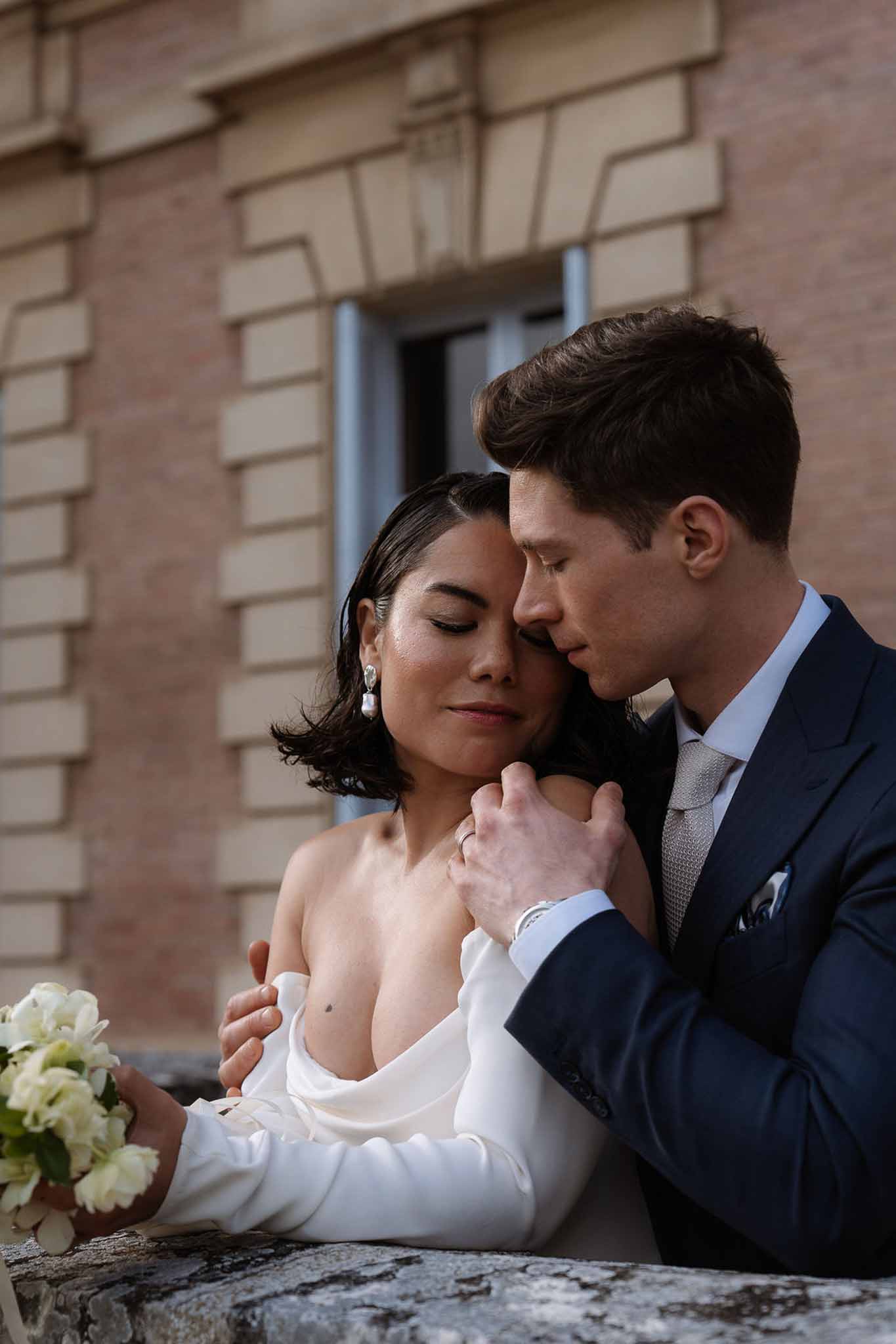 Bride in off-shoulder draped gown with white bouquet embraced by groom in navy suit against terracotta wall