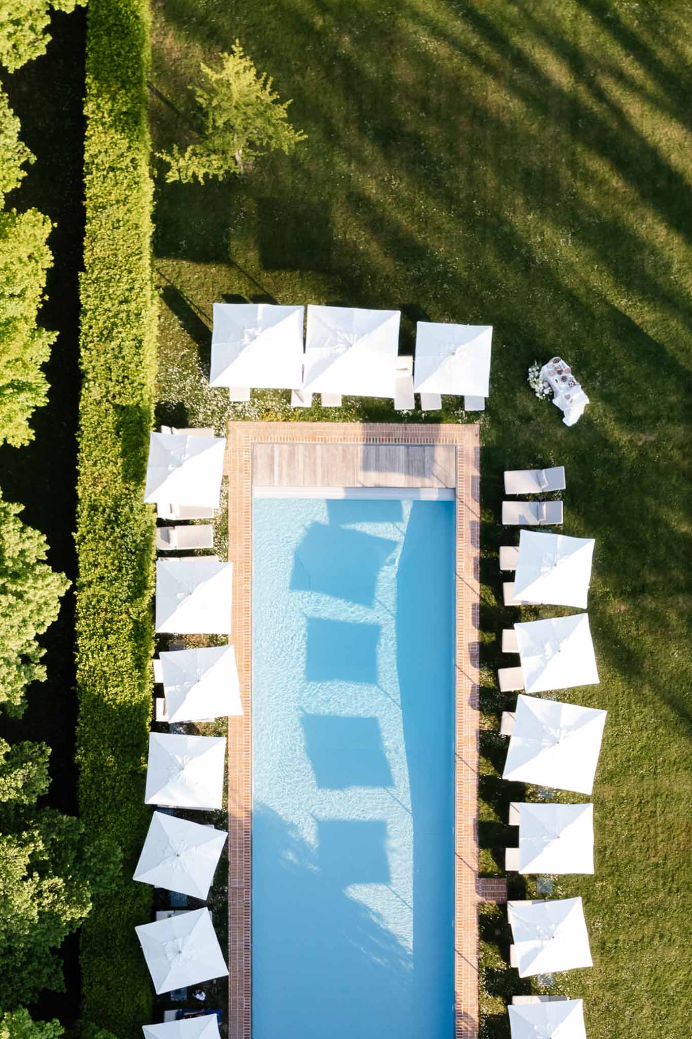 Aerial pool area with white market umbrellas, loungers, and wooden deck in formal hedged garden