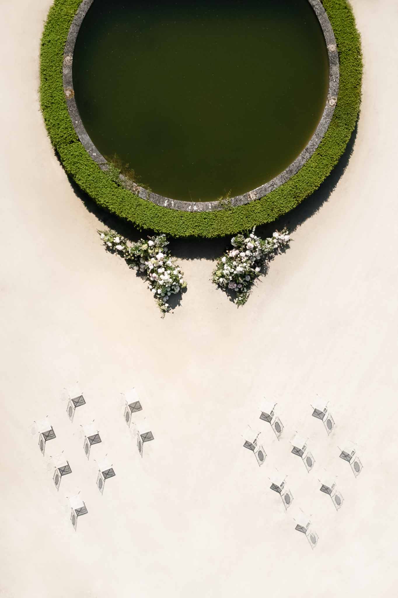 Aerial ceremony setup with white chairs around circular hedge pool and blush floral altar arrangements