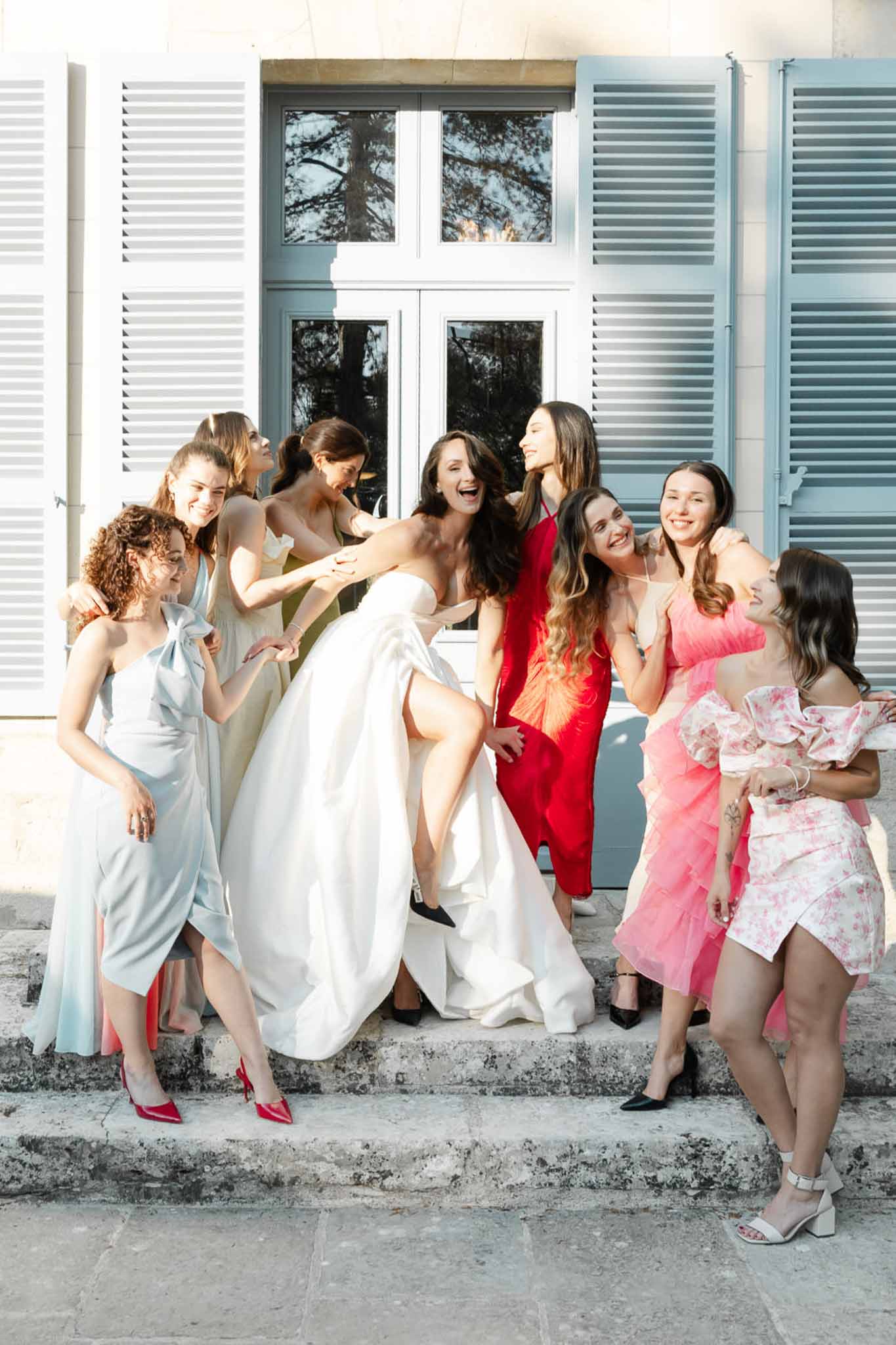 Bride in ballgown kicking leg up with seven mismatched bridesmaids laughing on stone steps