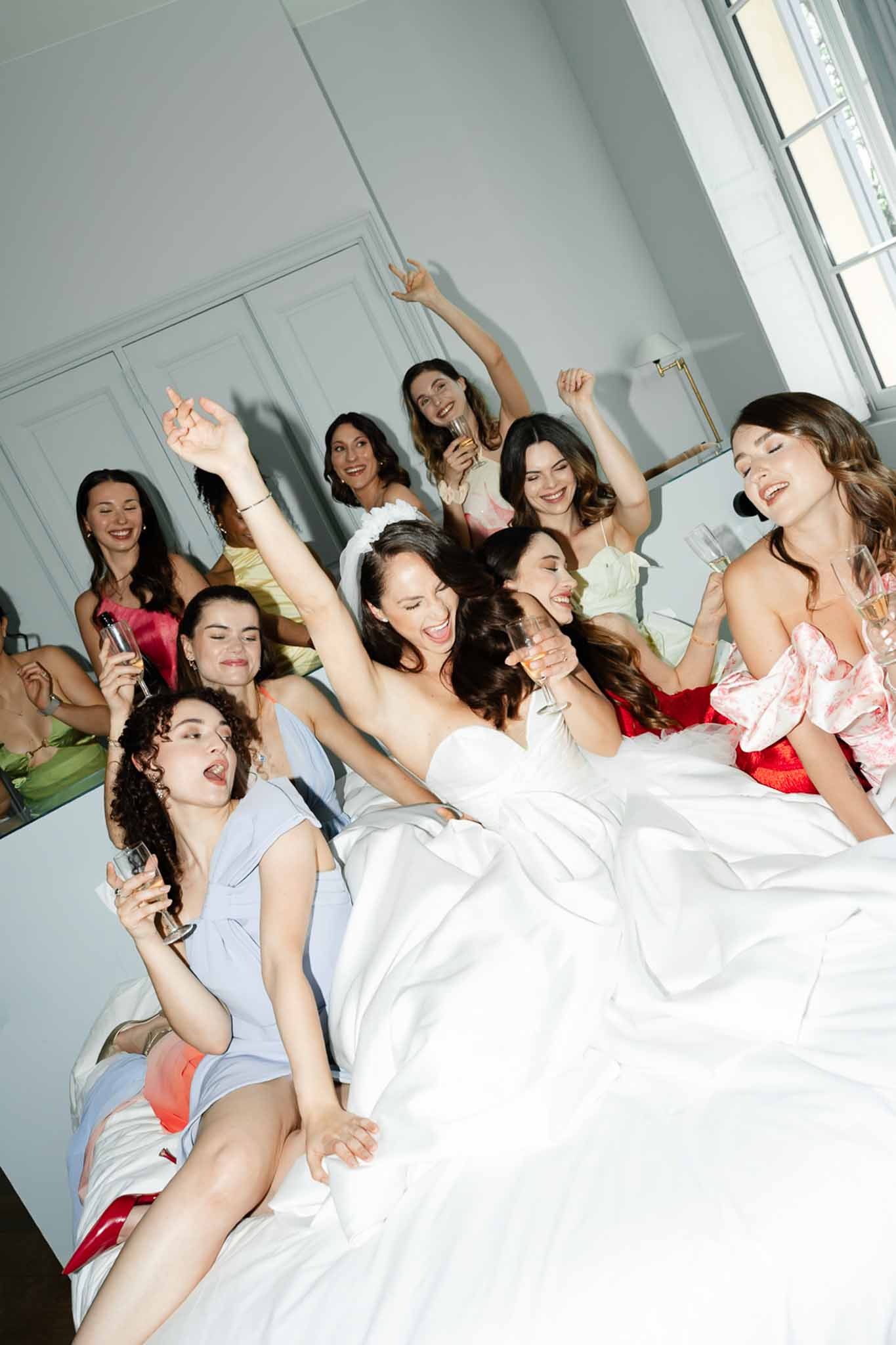 Bride and eight friends celebrate with champagne on bed in bright white-walled room before ceremony