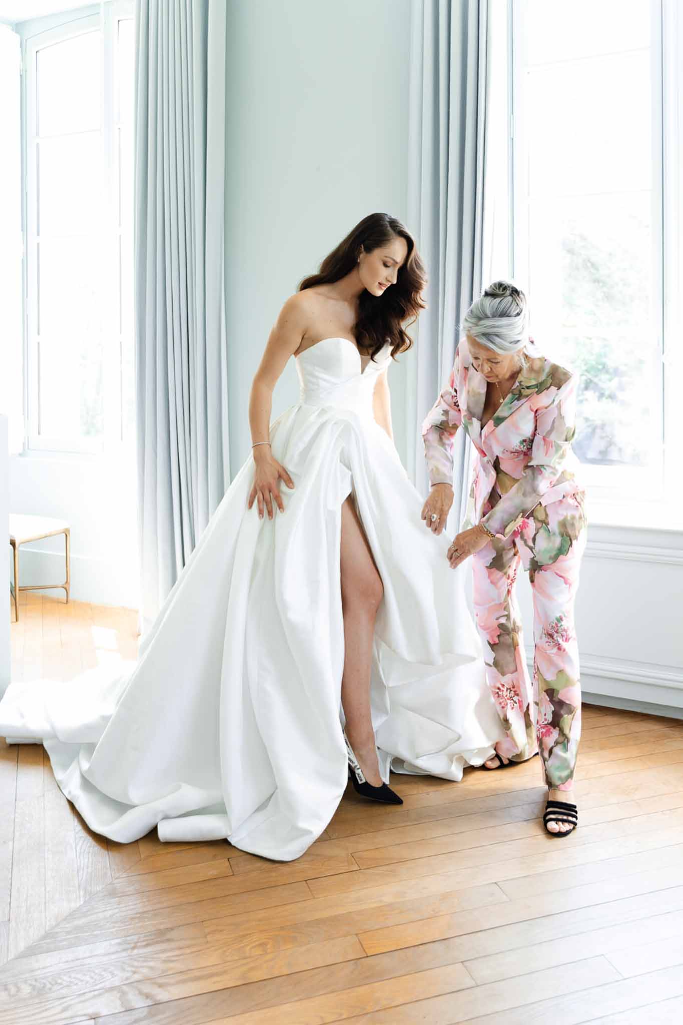 Bride and mother adjusting strapless ball gown train in pale blue chateau room with parquet floor