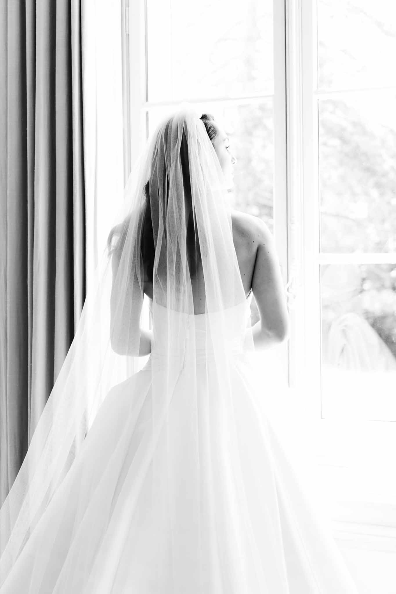 Black and white profile of bride in strapless ball gown with cathedral veil backlit at French window