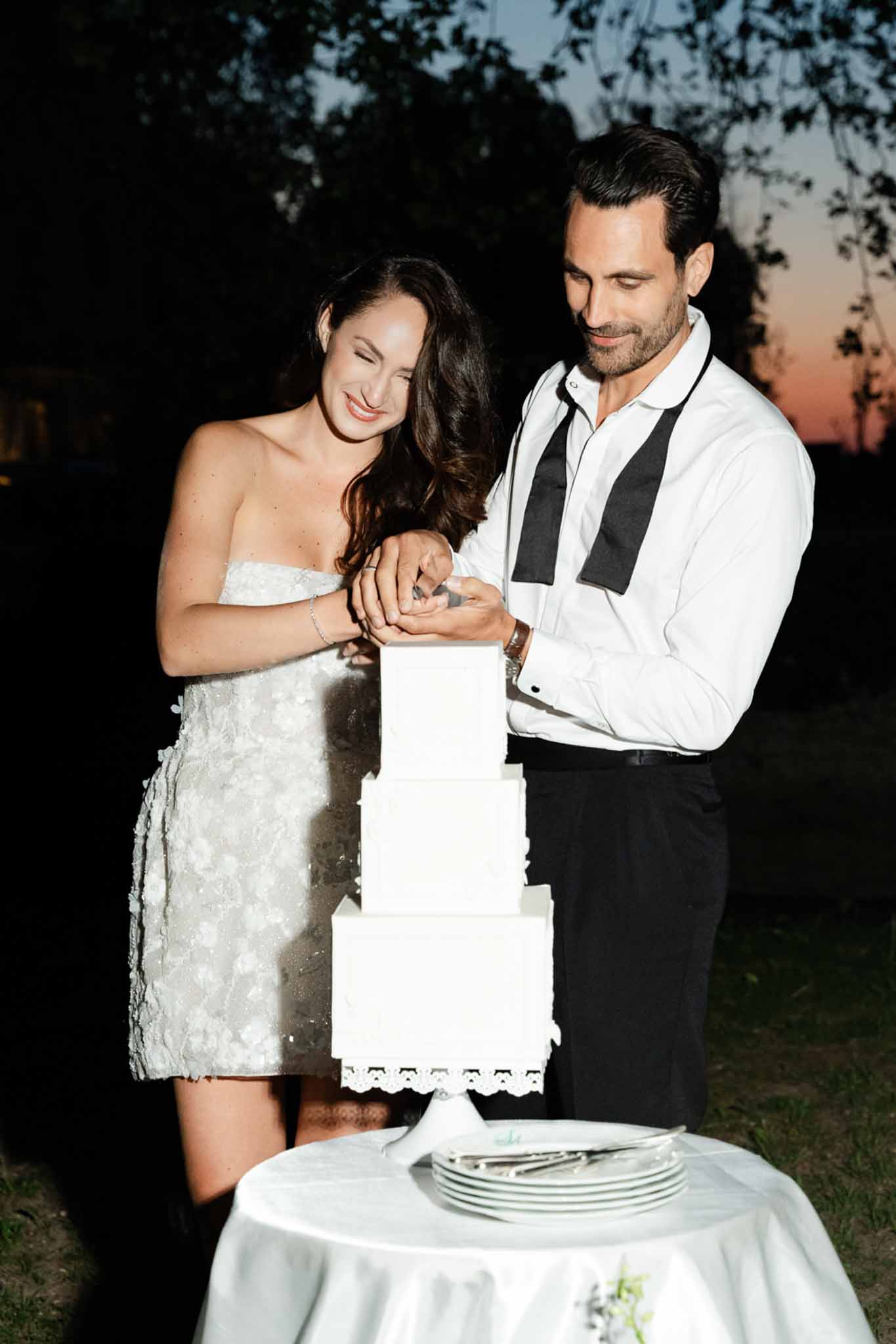 Couple cutting four-tier square white cake at dusk bride in textured mini dress groom with undone bow tie
