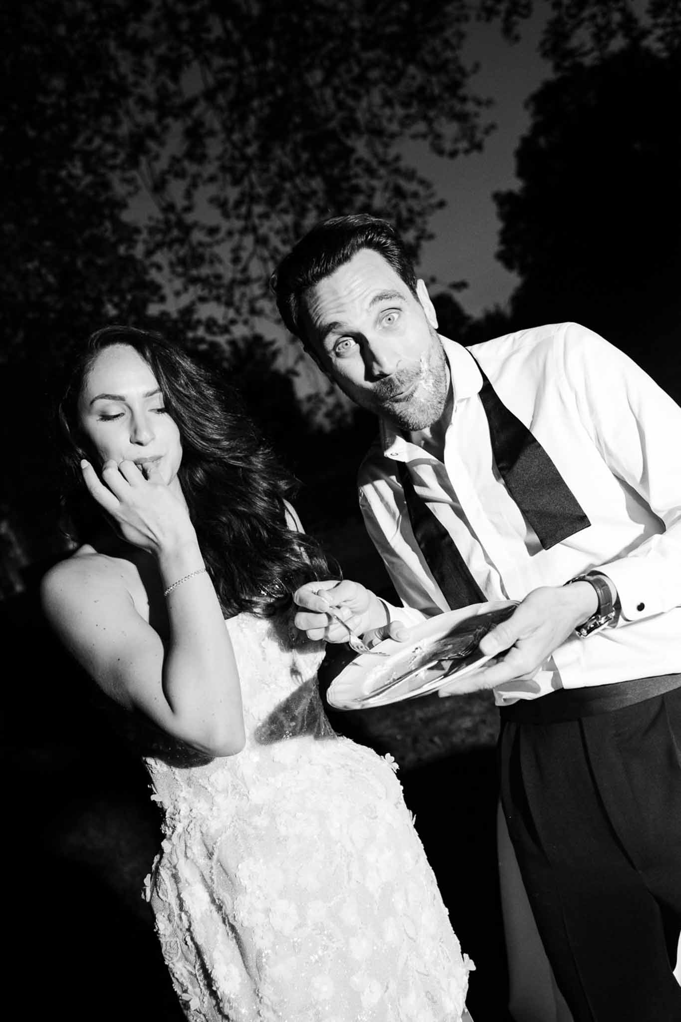 Groom with frosting on face and bride licking fingers during playful cake cutting in black and white