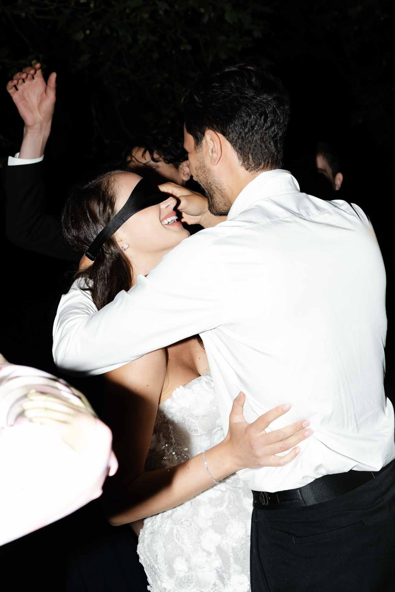 Bride in white lace gown wearing a blindfold laughing as groom leans in during a reception dance game