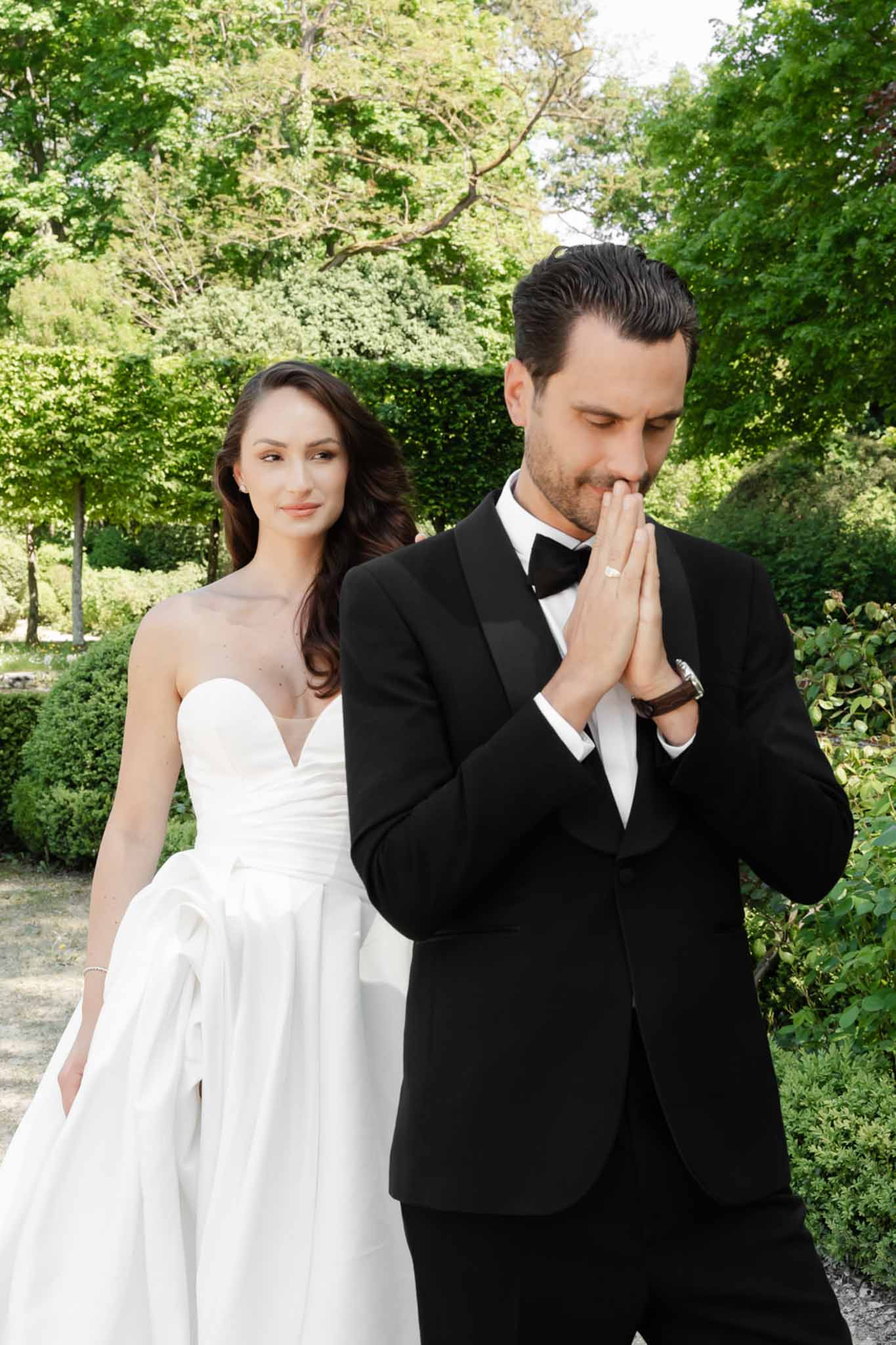Bride in strapless white ballgown and groom in black tuxedo posing candidly in manicured garden
