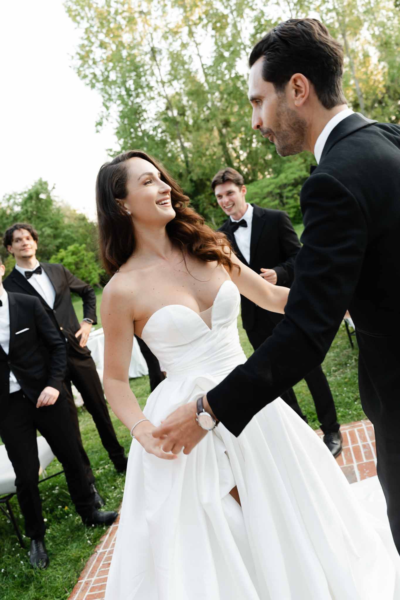 Groom holding laughing bride's hands on brick terrace with guests watching in background