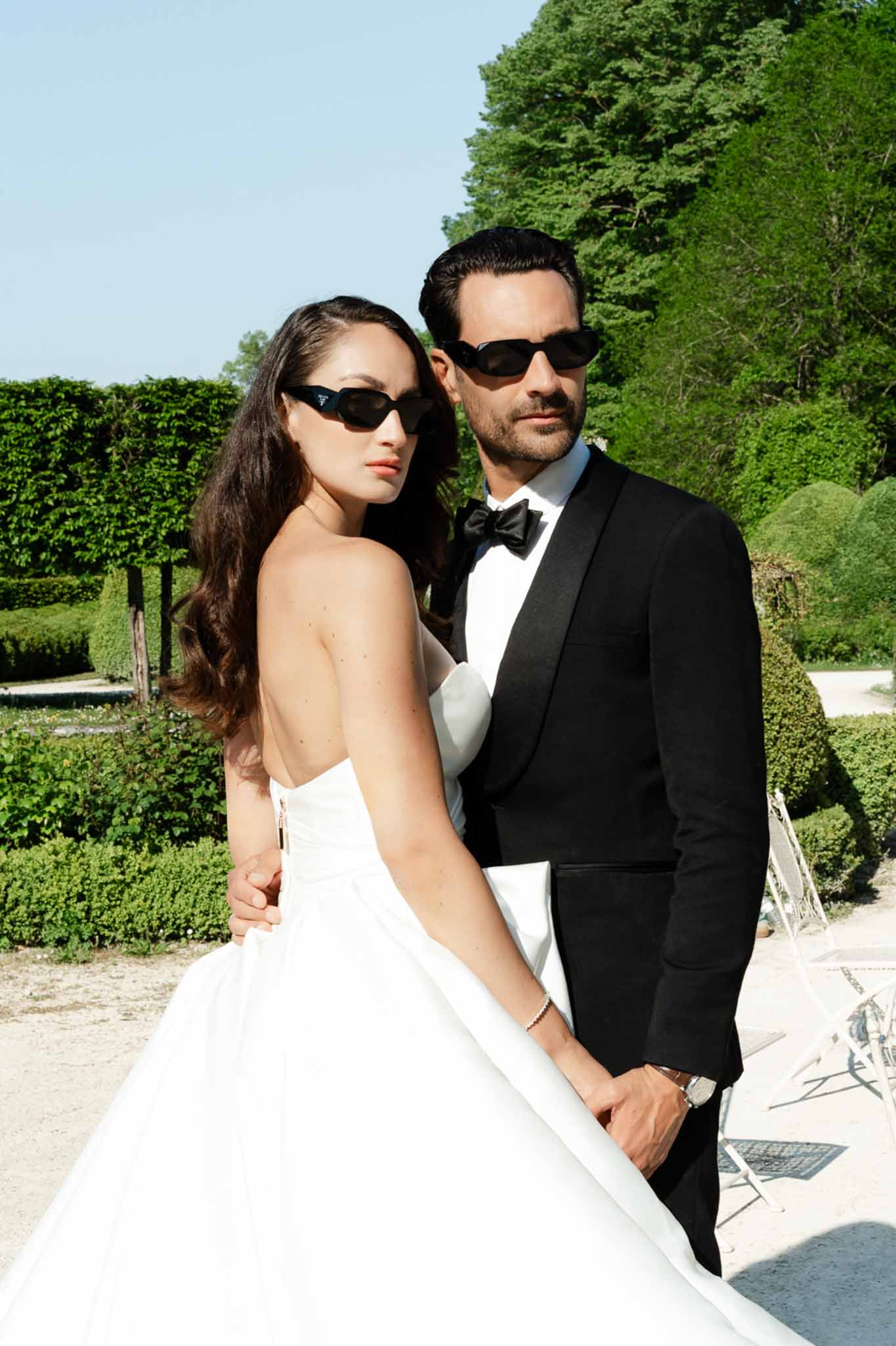 Couple in sunglasses faces camera with editorial pose in strapless ballgown and tuxedo in formal garden